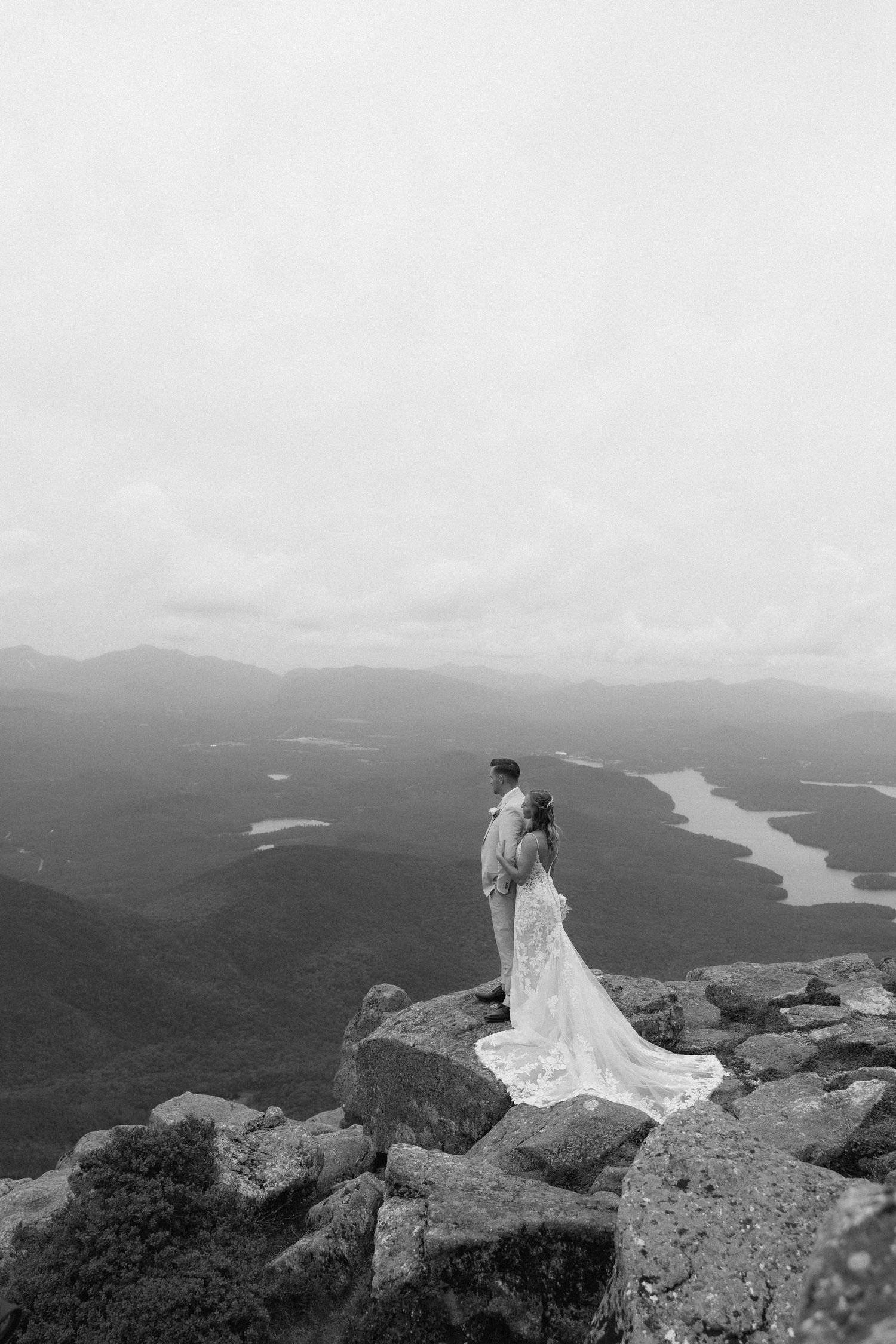 Bride and groom embrace on rocky mountain summit overlooking scenic valley and lakes in black and white wedding photo.