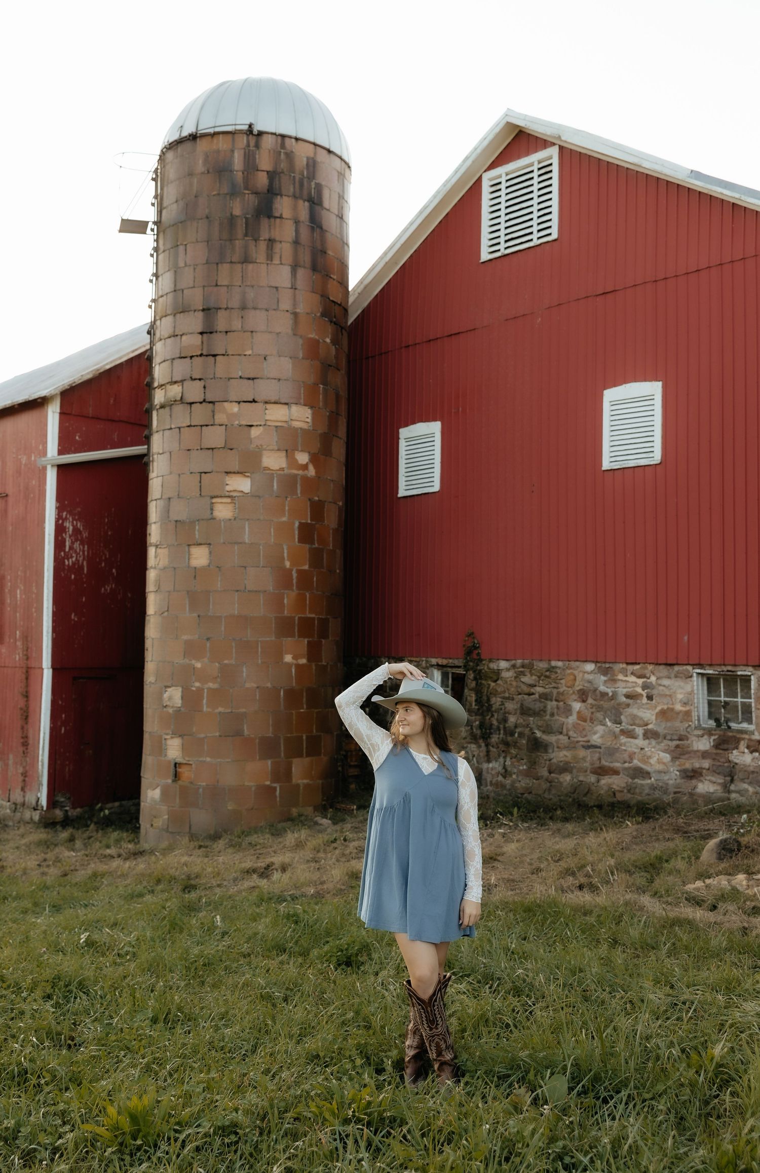 Woman in cowboy hat and blue dress poses by rustic brick silo and red barn on a farm.