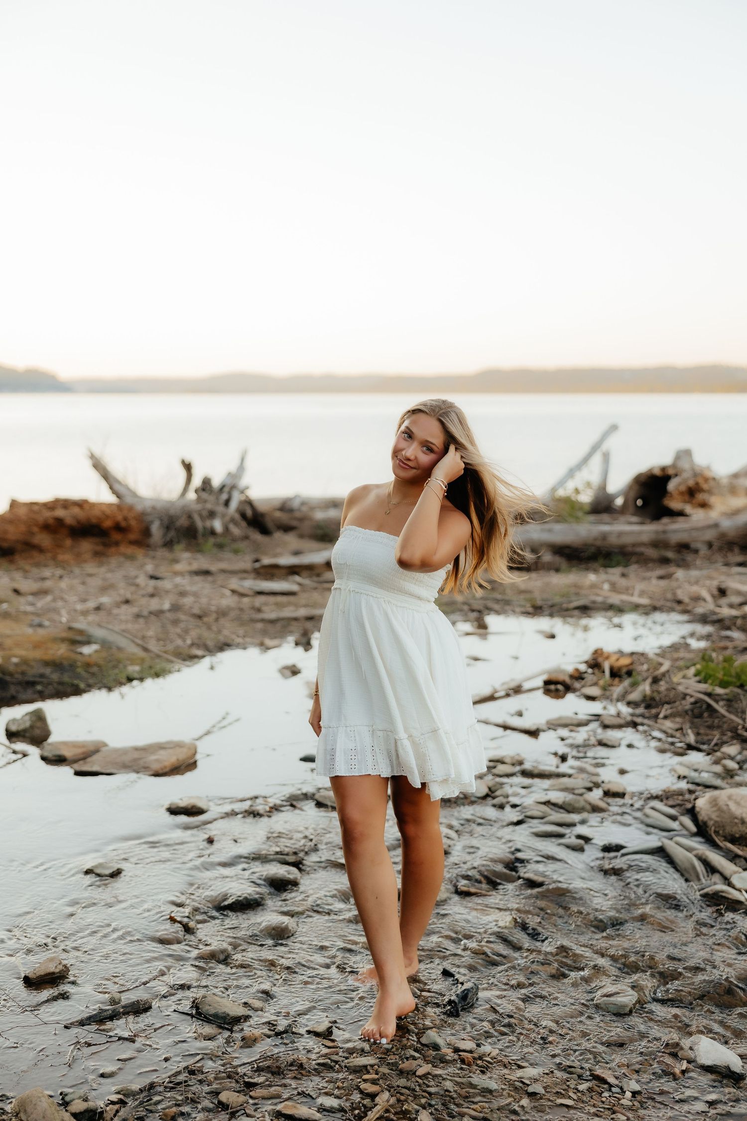 Young woman in white sundress standing barefoot on rocky beach shore at sunset with driftwood background.