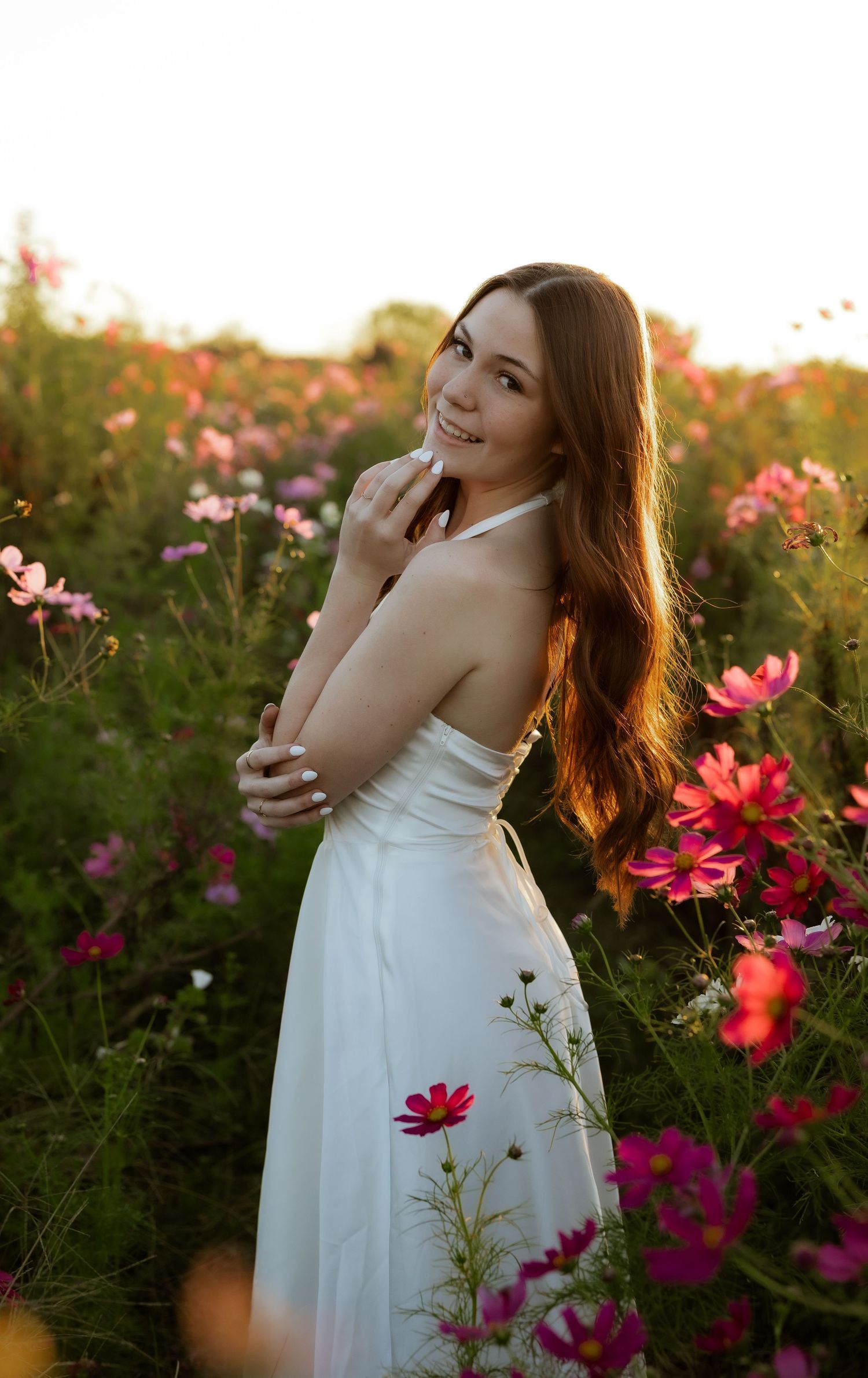 A dreamy scene in a cosmos flower field at golden hour with a white sundress and warm sunlight filtering through.