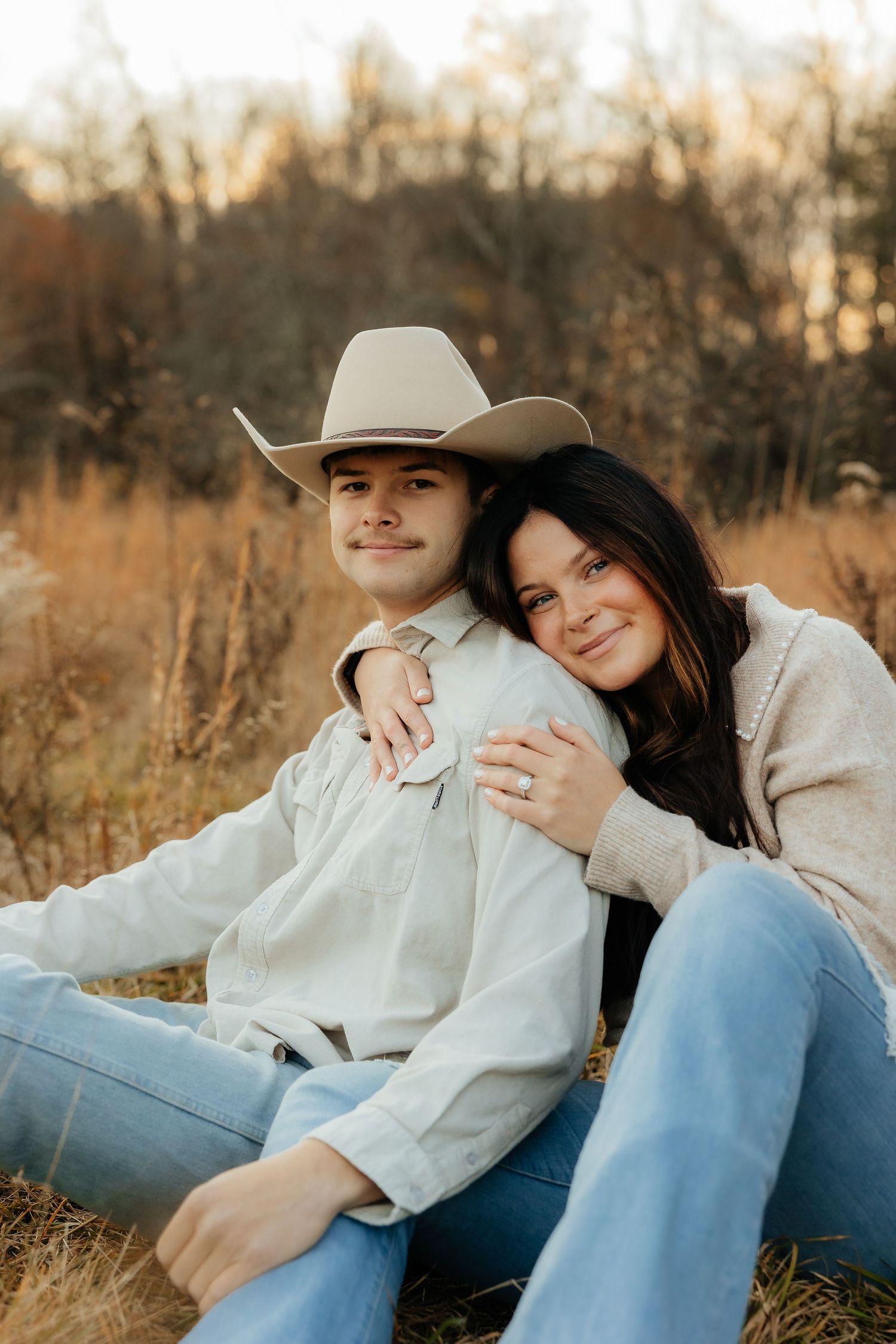 A couple in western attire sits together in a field during golden hour, embracing in casual denim and cozy sweaters.