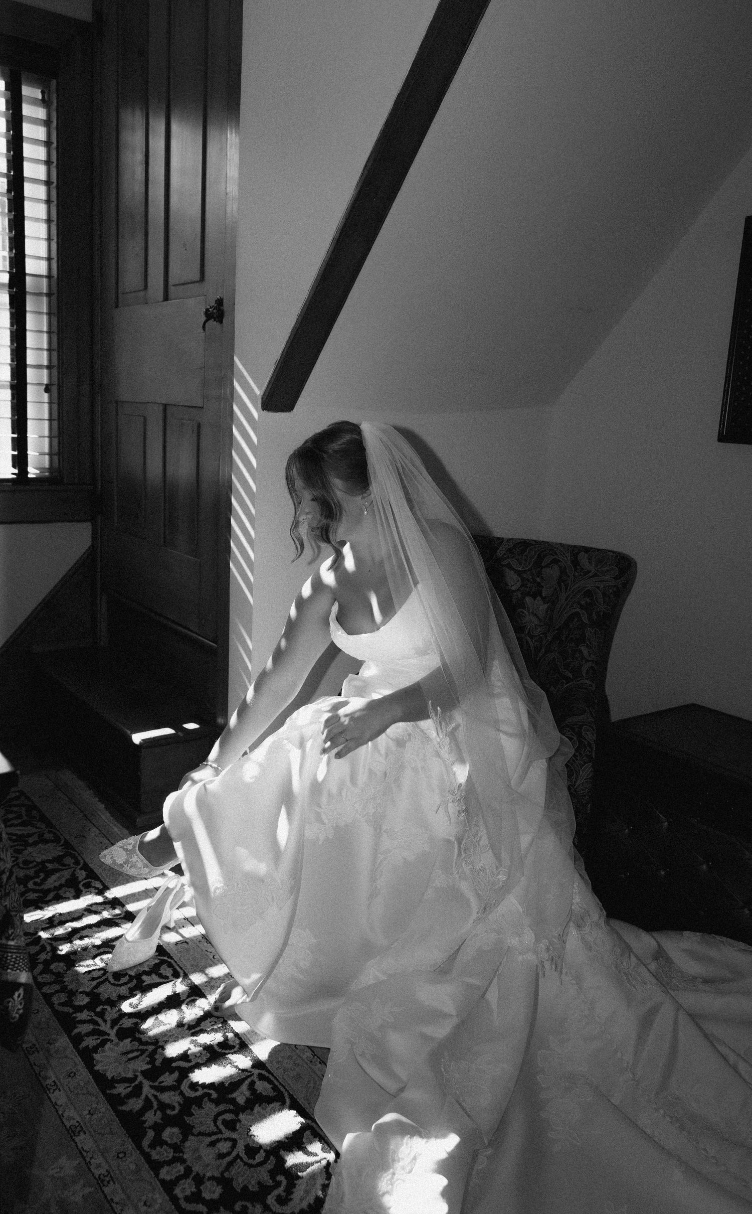 Black and white photo of a bride in a white gown and veil sitting by a window with light streaming in.