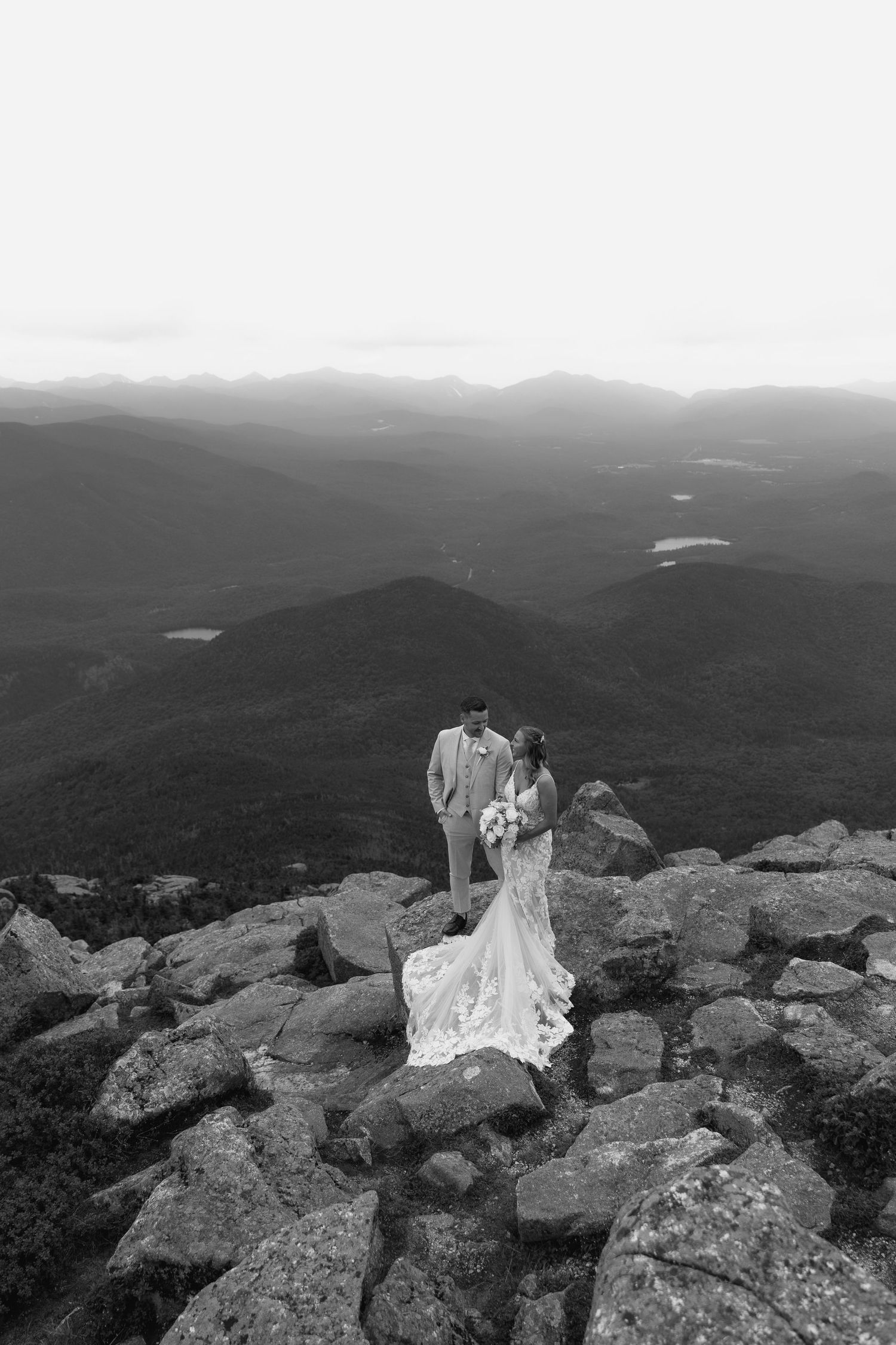 Black and white photo of bride and groom standing on rocky mountain summit with sweeping valley views.