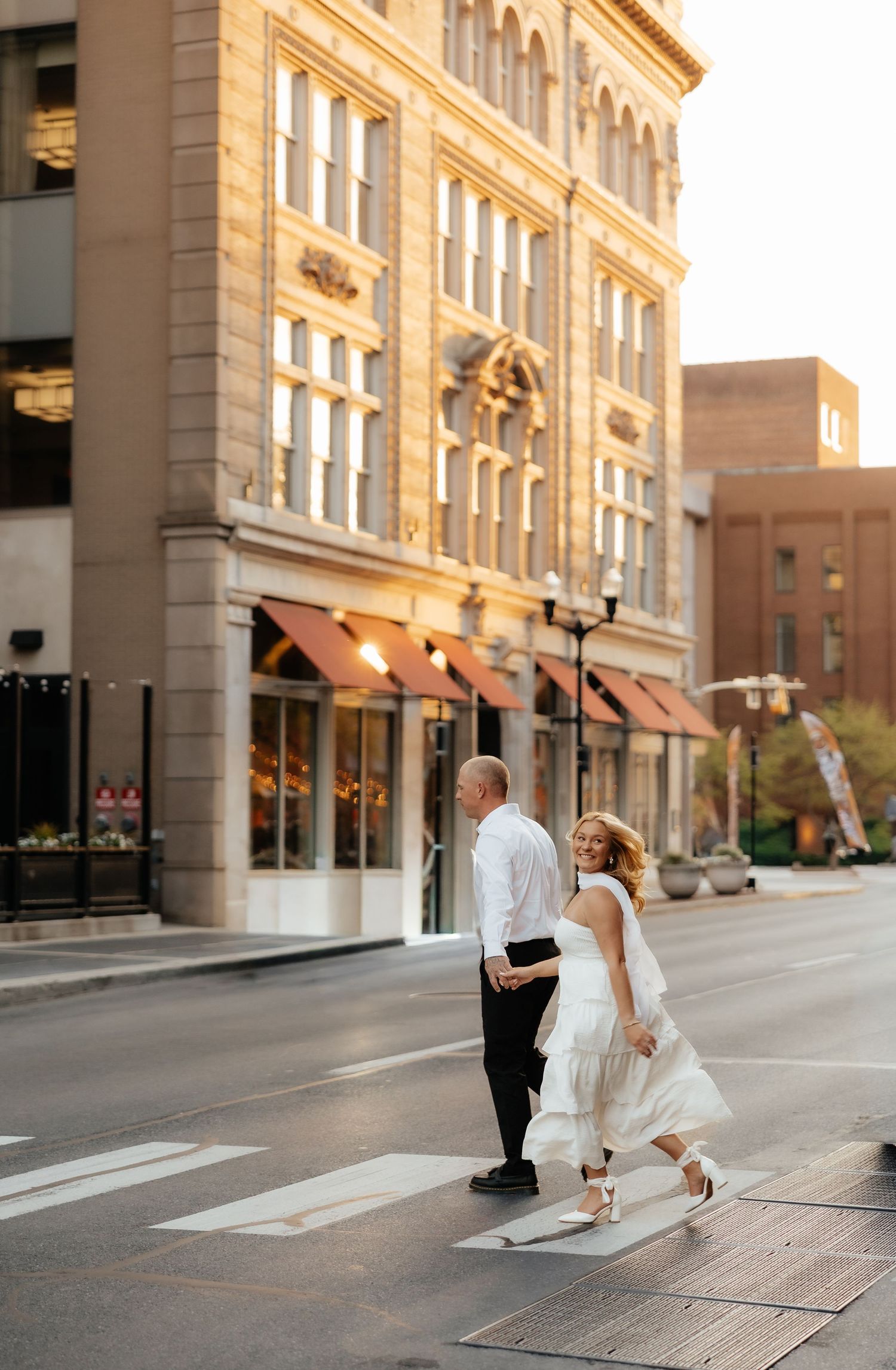 Couple in wedding attire crossing a downtown street at golden hour near a historic brick building.