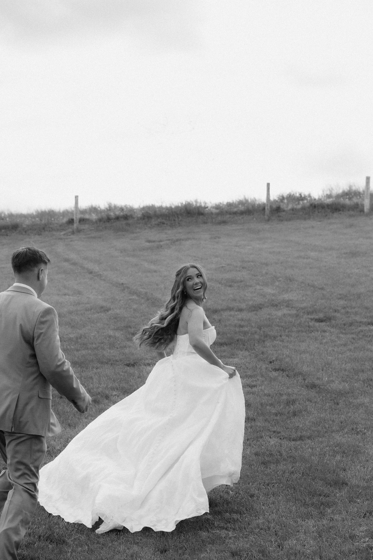 Black and white photo of a laughing bride in a flowing gown holding hands with groom walking through a green field.