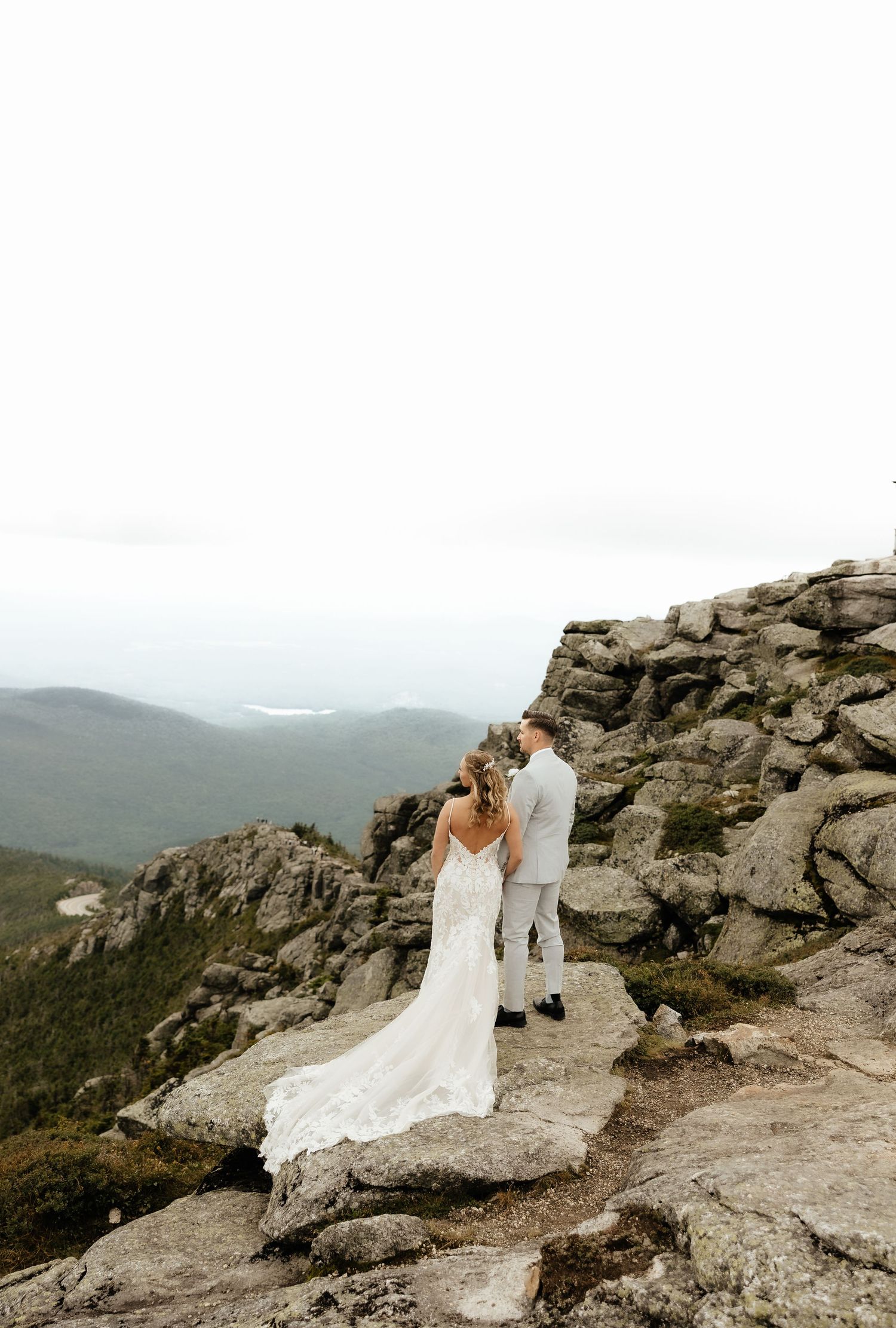 Bride and groom stand on rocky mountain summit in wedding attire, overlooking misty valley landscape.