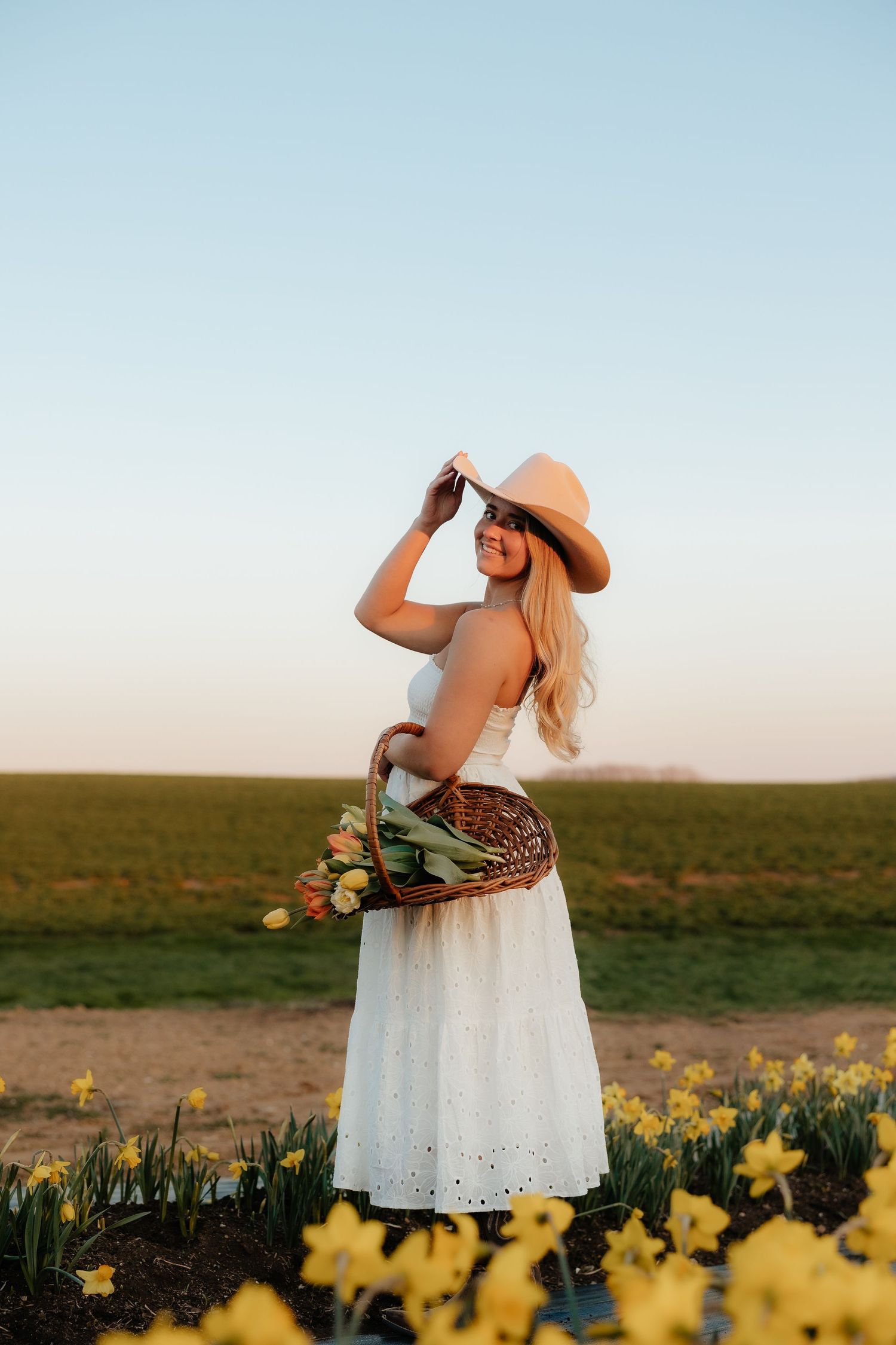 Woman in white dress and straw hat holding a wicker basket of tulips in a sunny flower field.