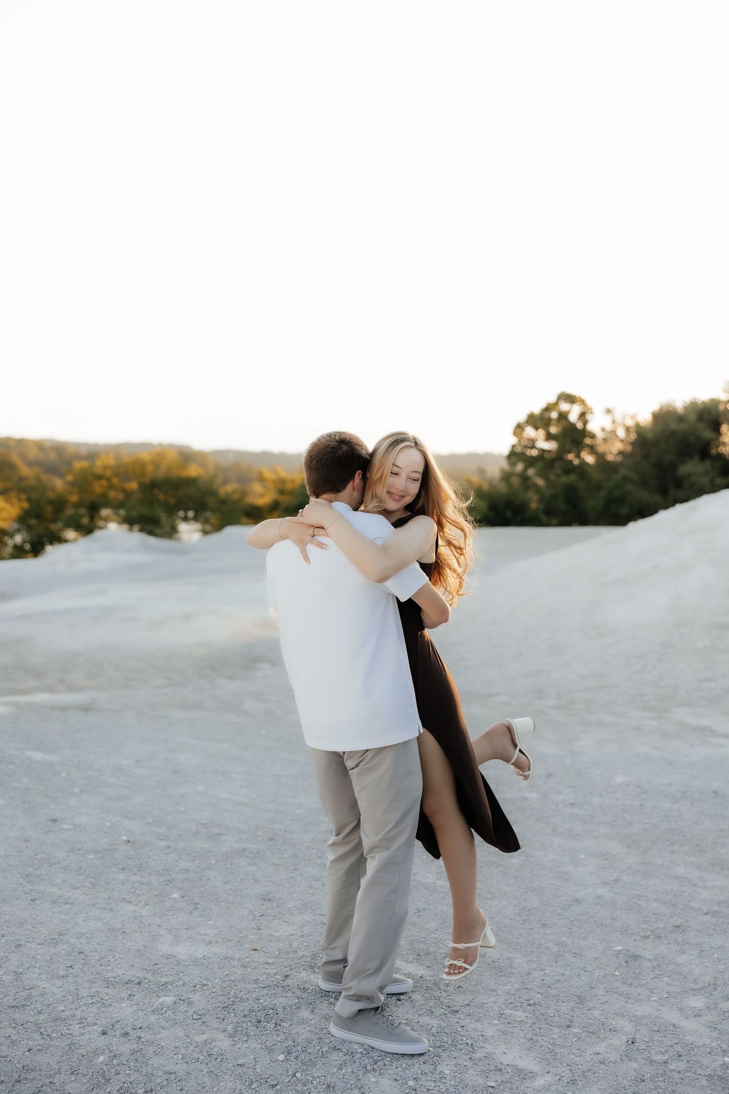 A joyful couple embraces outdoors, woman in black dress laughing as man lifts her on a rocky landscape at golden hour.