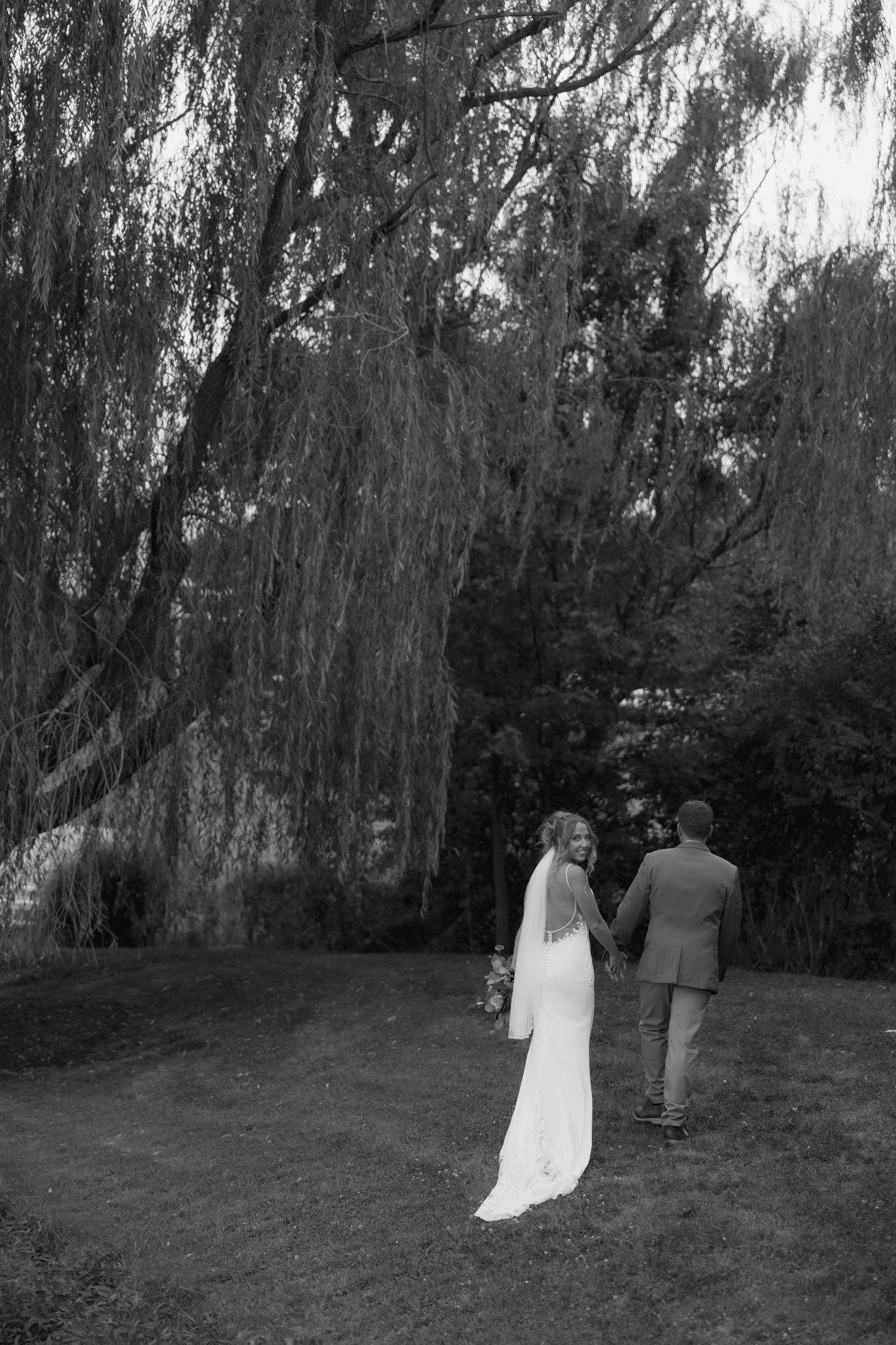 Black and white photo of bride and groom walking hand in hand beneath a weeping willow tree on their wedding day.
