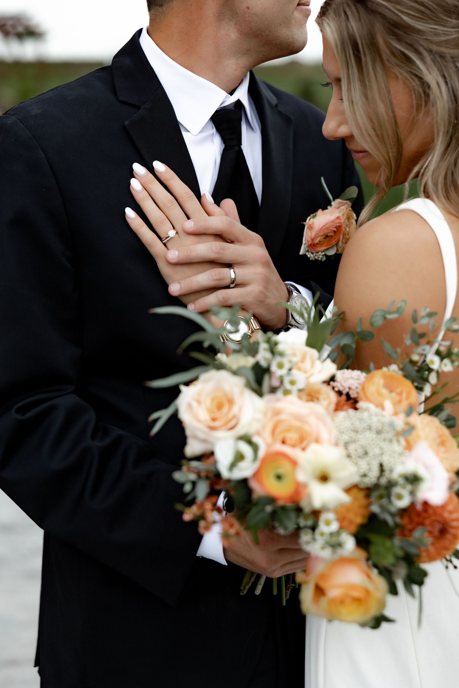 Close-up wedding photo showing hands with rings and a peachy orange floral bouquet with white accents.