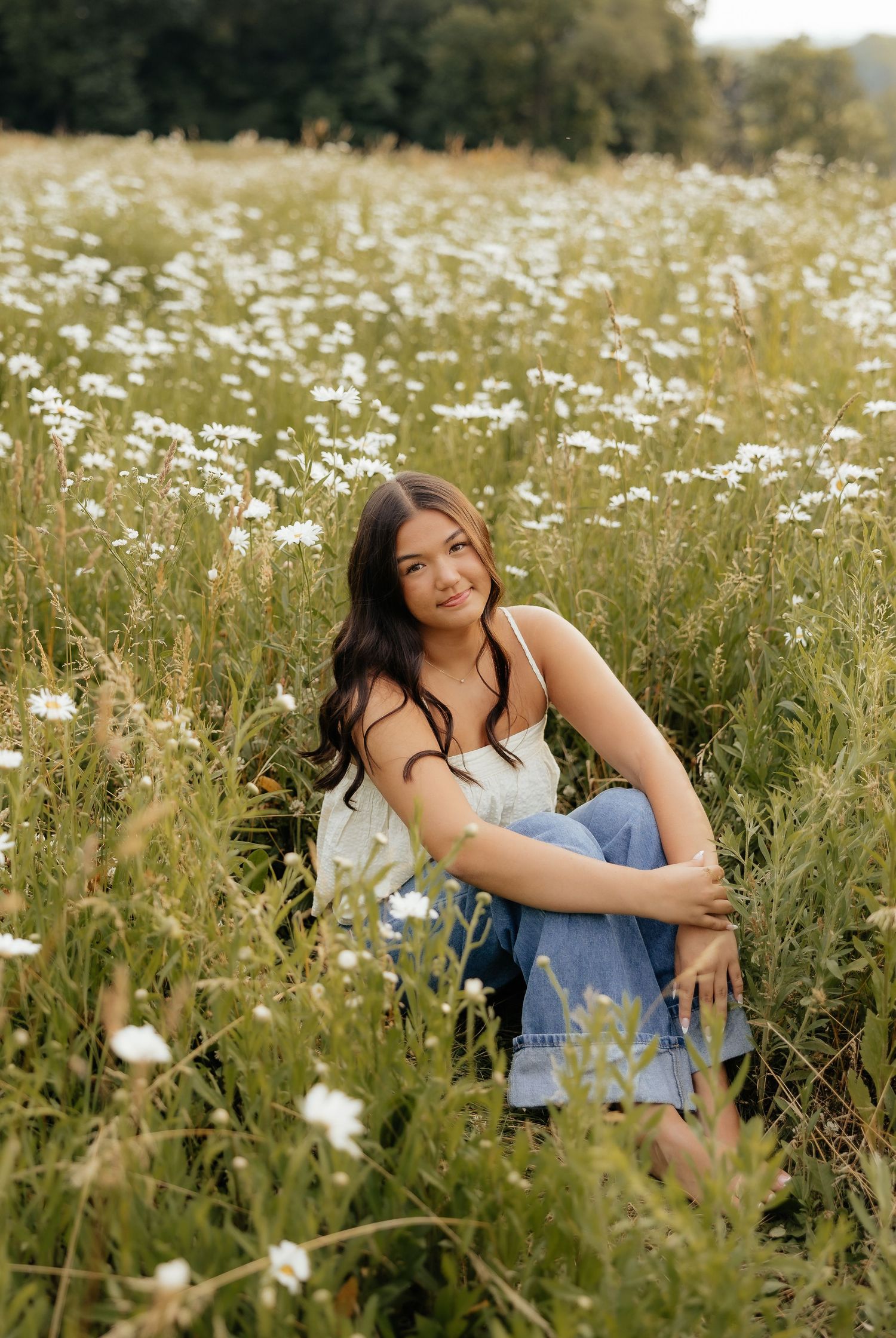Young woman sitting in a wildflower field wearing a white top and jeans, surrounded by white daisies.