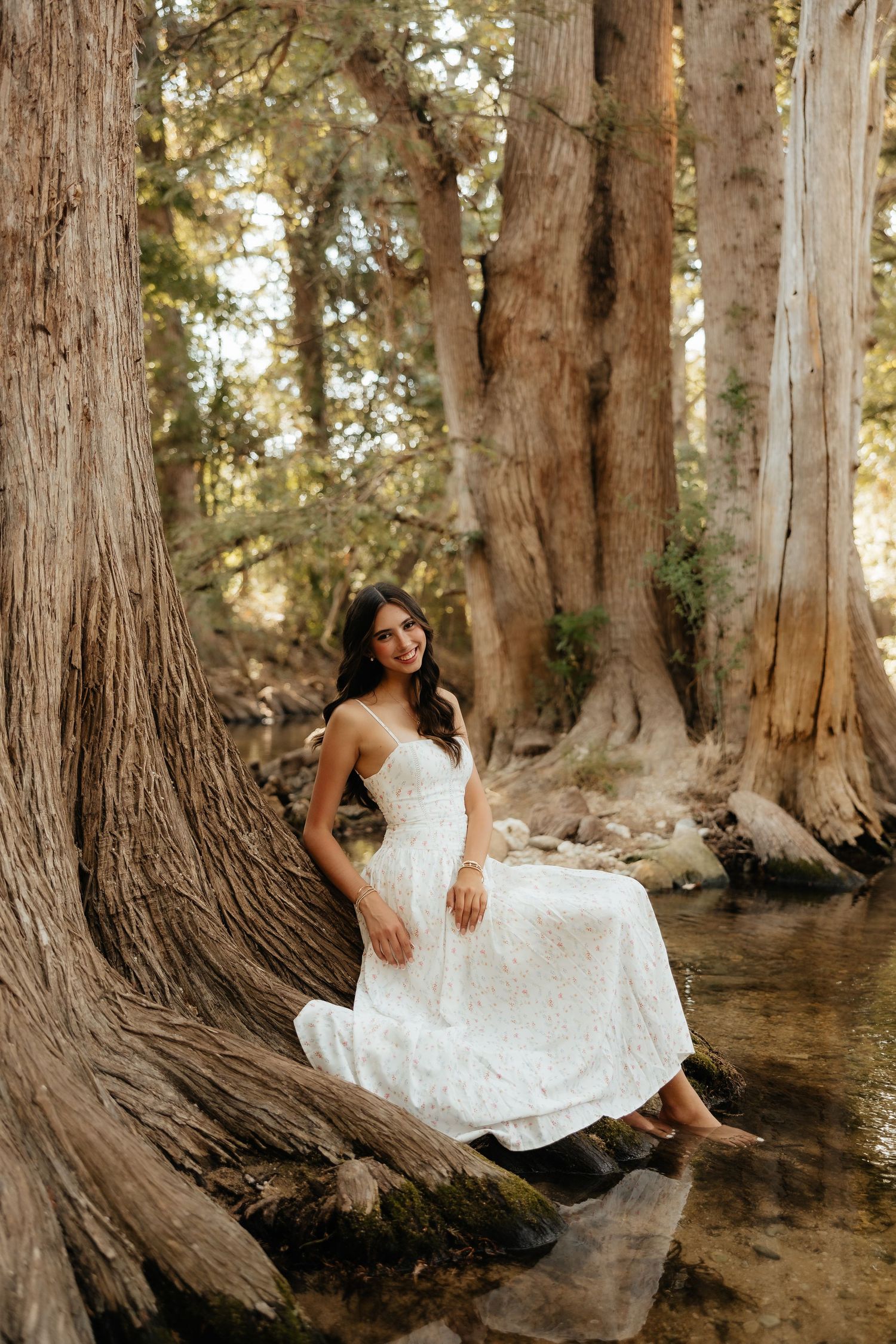 A bride in a white lace gown sits gracefully on rocks by a stream surrounded by tall cypress trees.