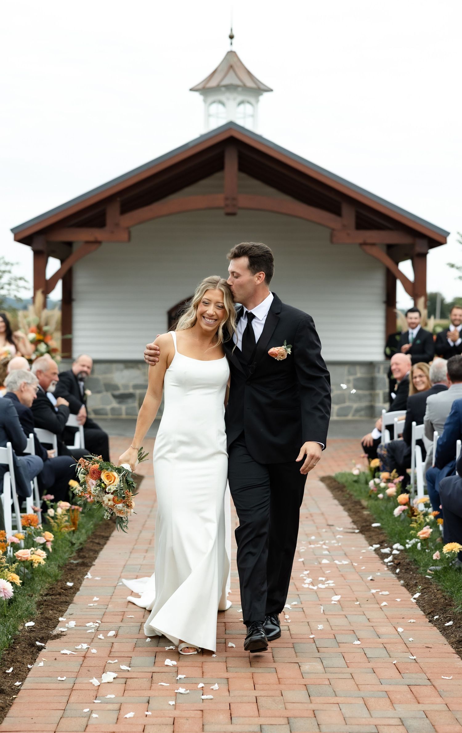 Wedding couple walks along brick pathway lined with flowers outside white chapel building.