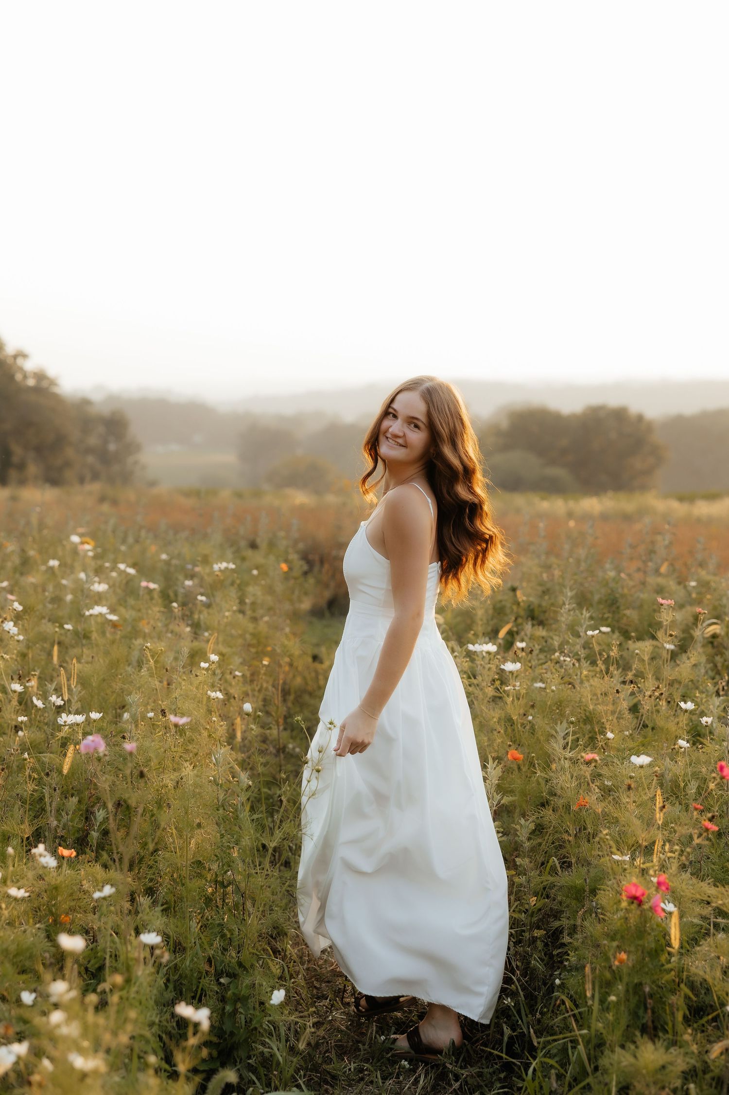 Young woman in white dress standing in a wildflower field at golden hour, smiling over her shoulder.