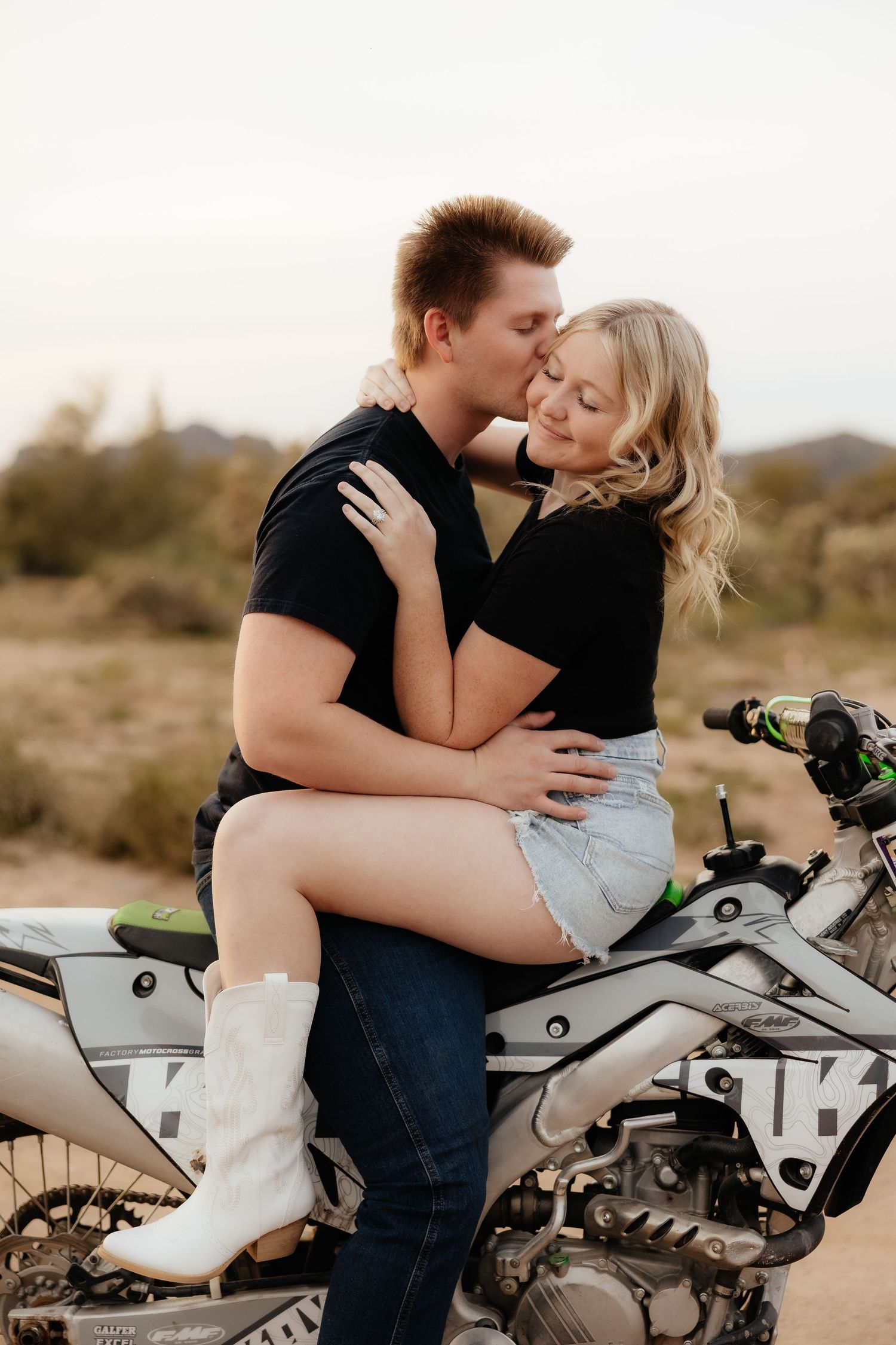 A romantic couple shares intimate moments on a dirt bike in a desert landscape during a photoshoot.