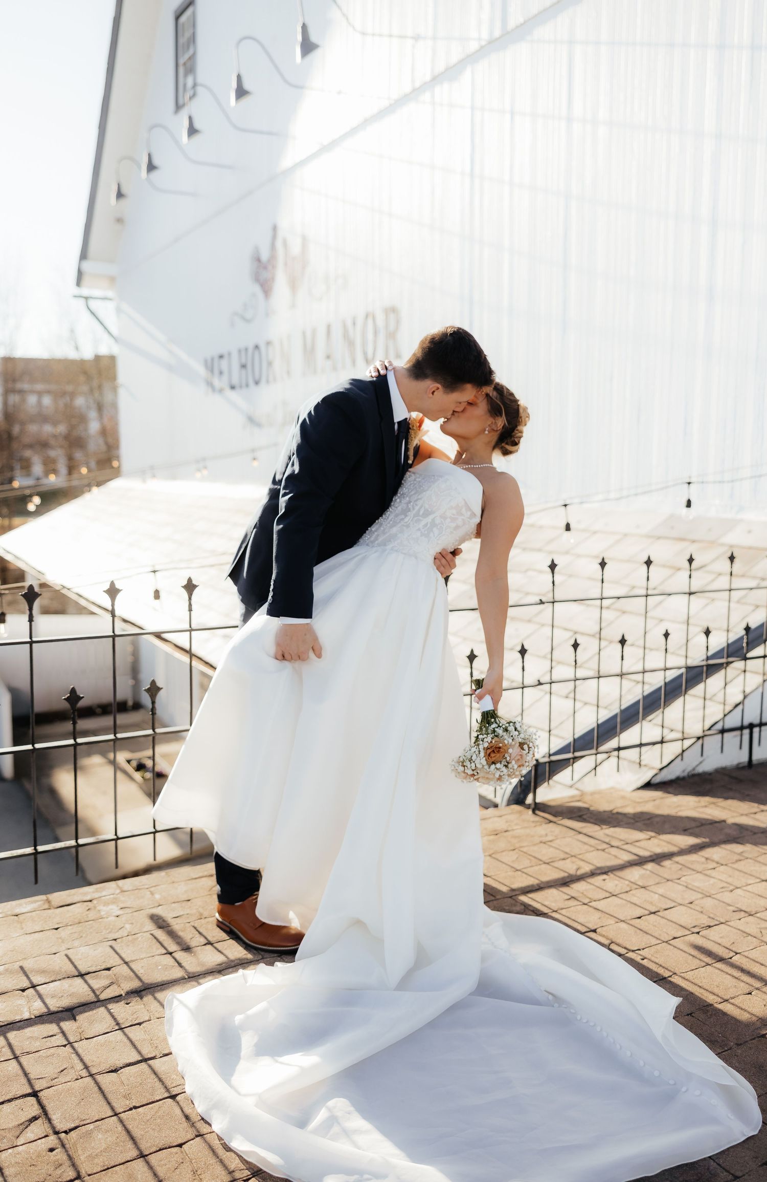 Bride and groom sharing a romantic kiss outdoors at Melhorn Manor, bride in white gown holding bouquet.