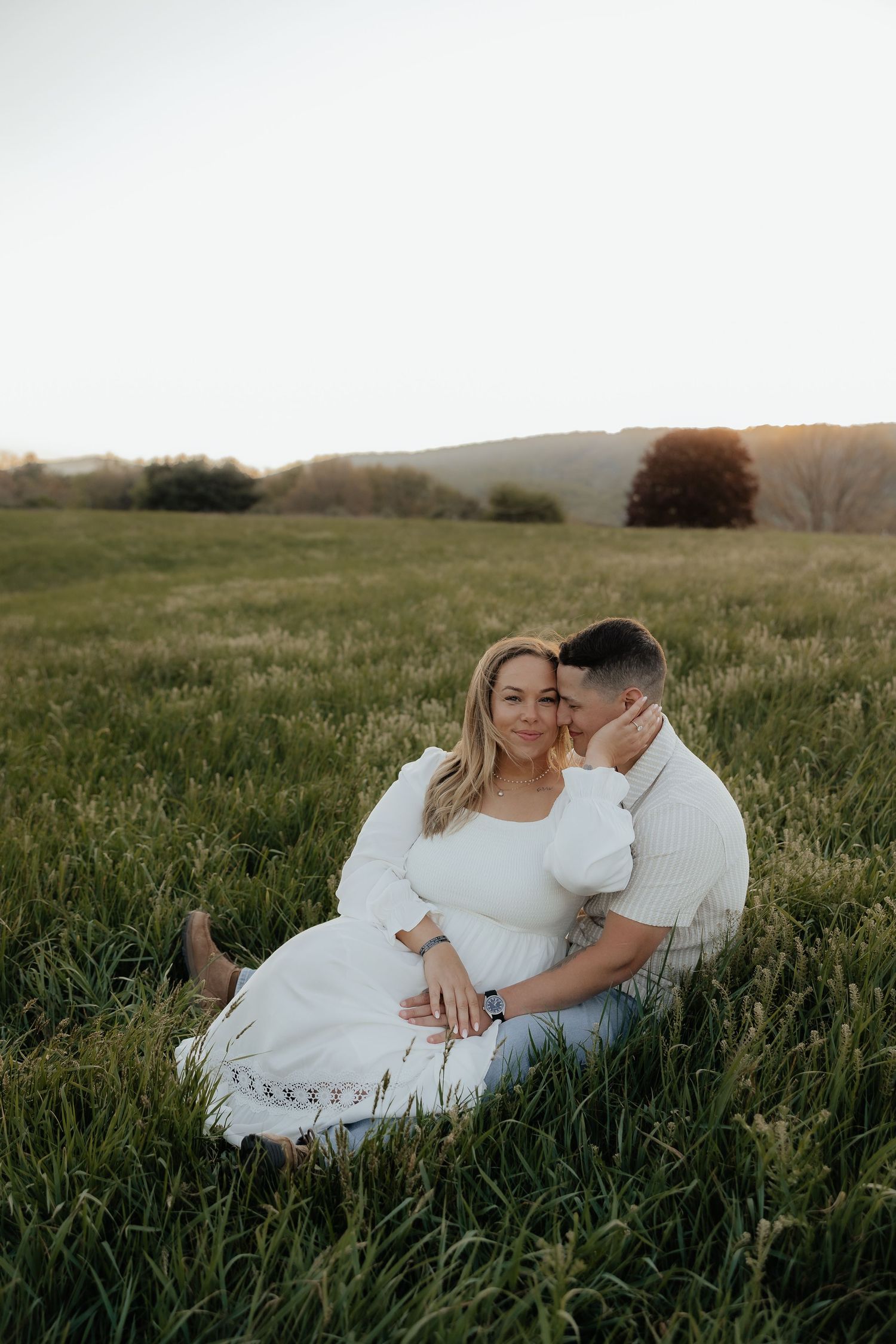 Couple sitting in lush green field at sunset, woman in white dress leaning against man in white sweater.