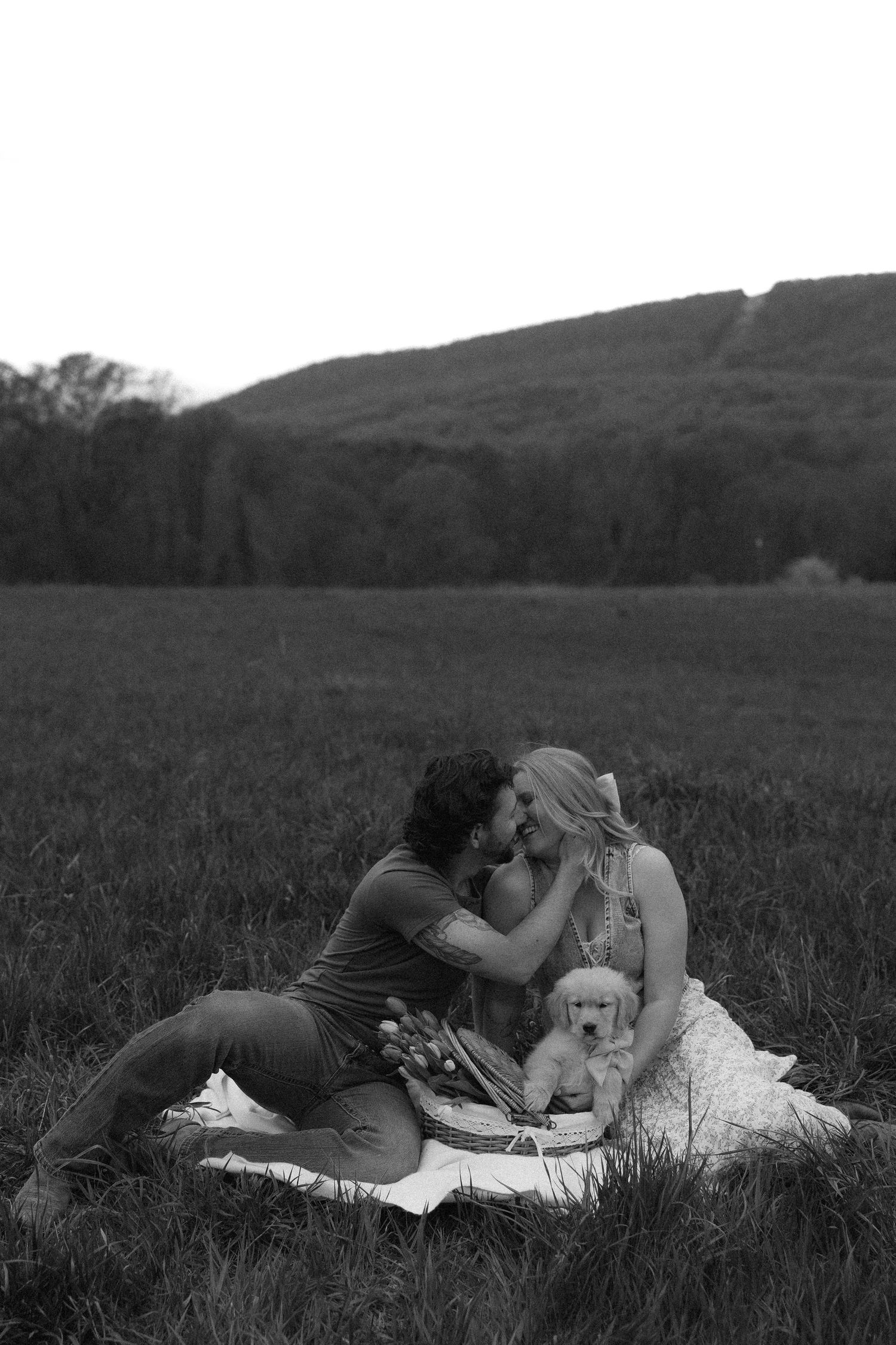 Black and white photo of a couple kissing on a blanket in a field with a puppy and mountains in the background.