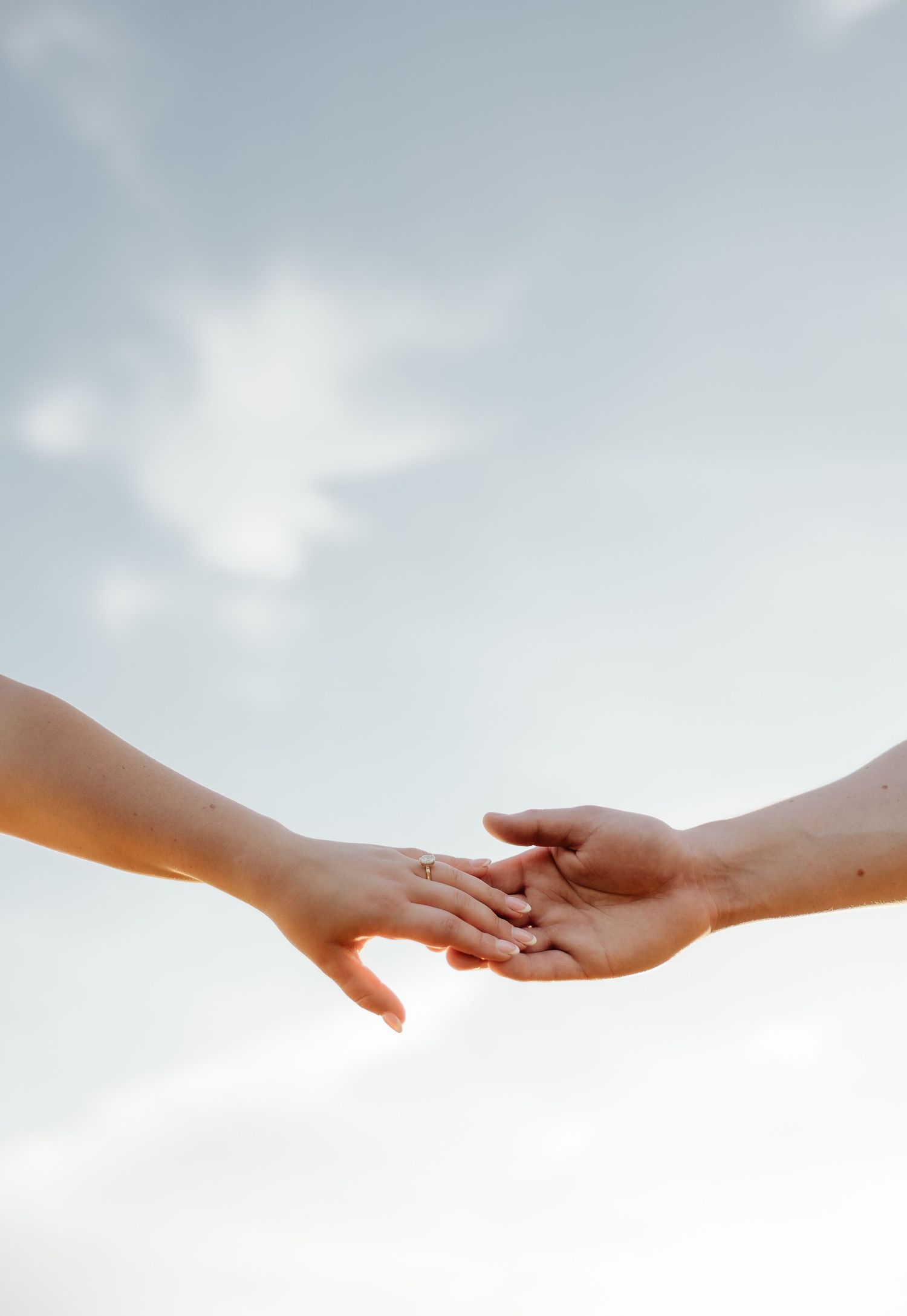 Two hands gently reaching toward each other against a soft cloudy sky, one adorned with a delicate ring.