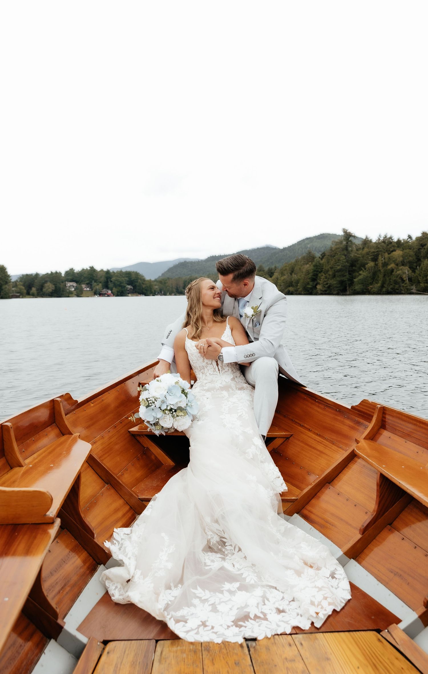 Bride and groom share a romantic moment in a wooden rowboat on a lake, bride holding white bouquet in lace gown.