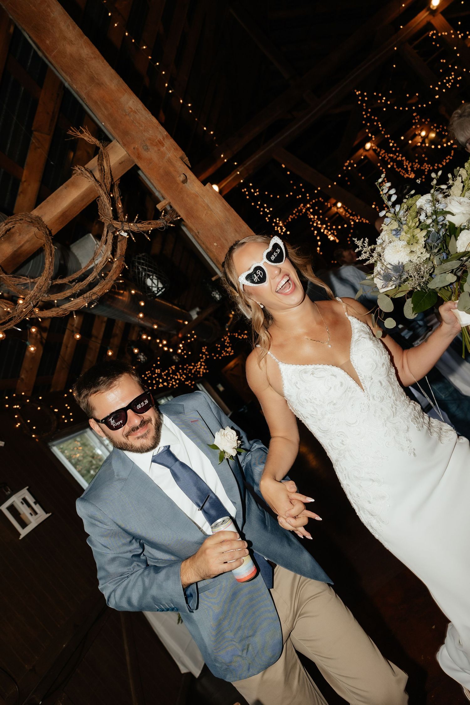 Bride and groom wearing fun sunglasses celebrate at rustic barn wedding reception with string lights.