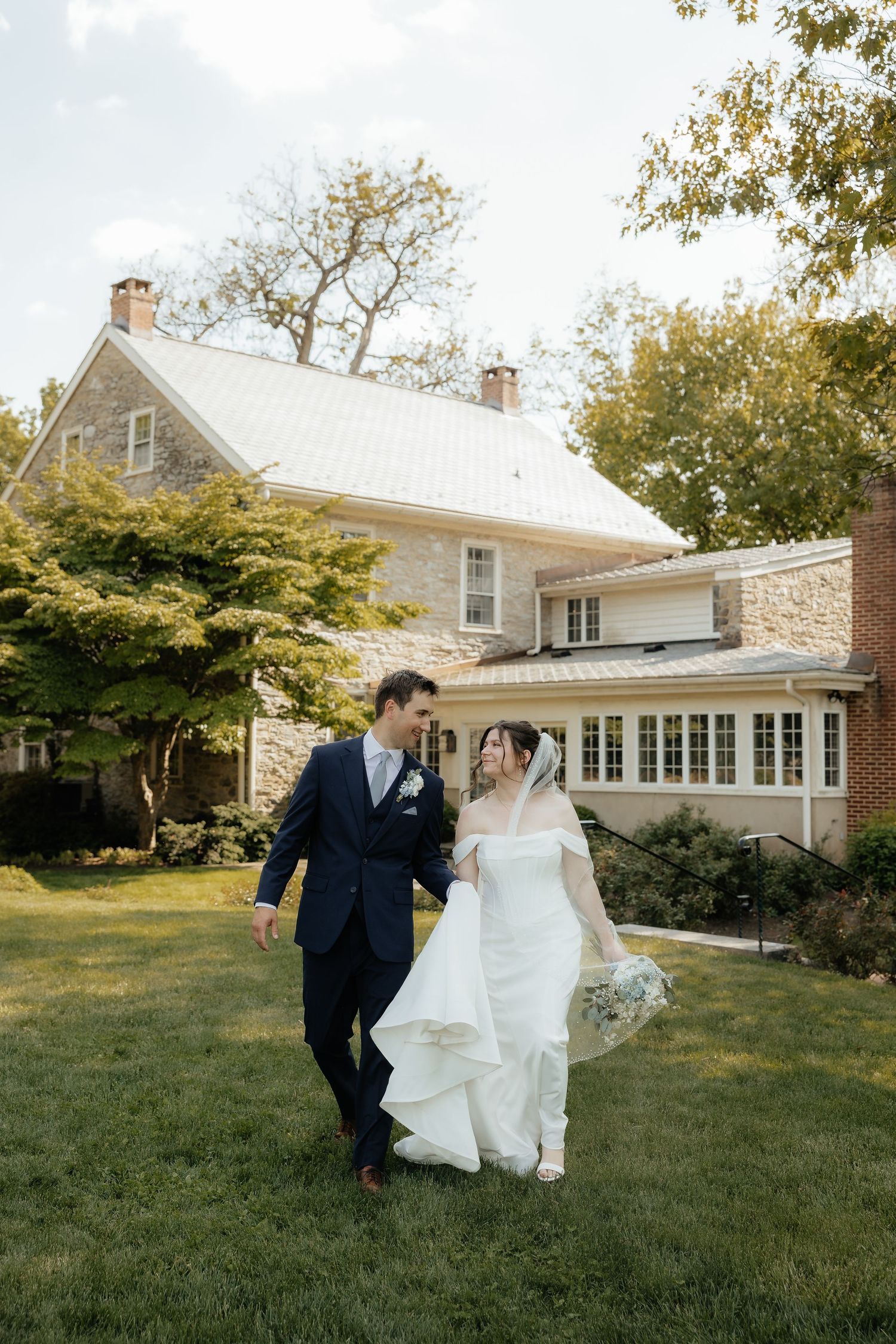 Bride and groom walk hand-in-hand on lush green lawn outside elegant stone manor house on their wedding day.