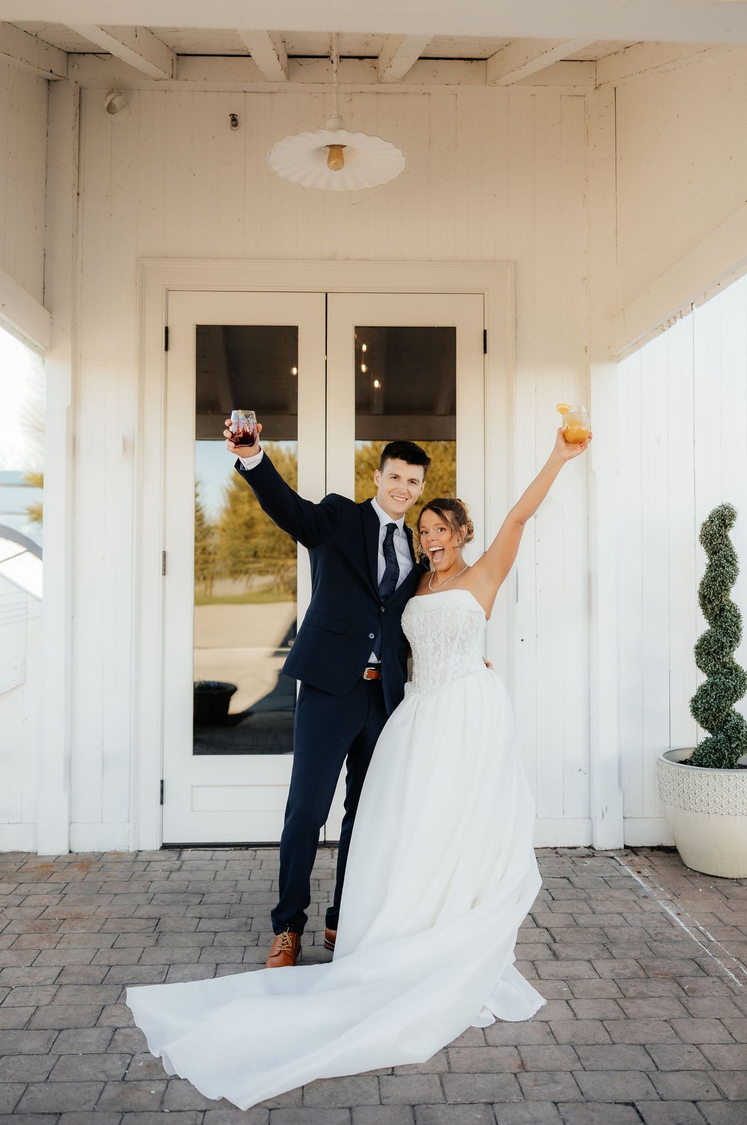 Joyful newlyweds celebrate outdoors, raising cocktails in front of white doors, bride in strapless gown, groom in navy suit.