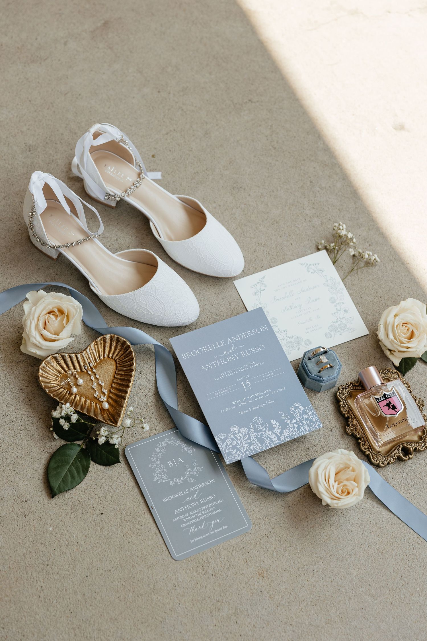 Elegant wedding flat lay featuring white bridal shoes, dusty blue invitations, cream roses, perfume, and gold heart trinket dish.