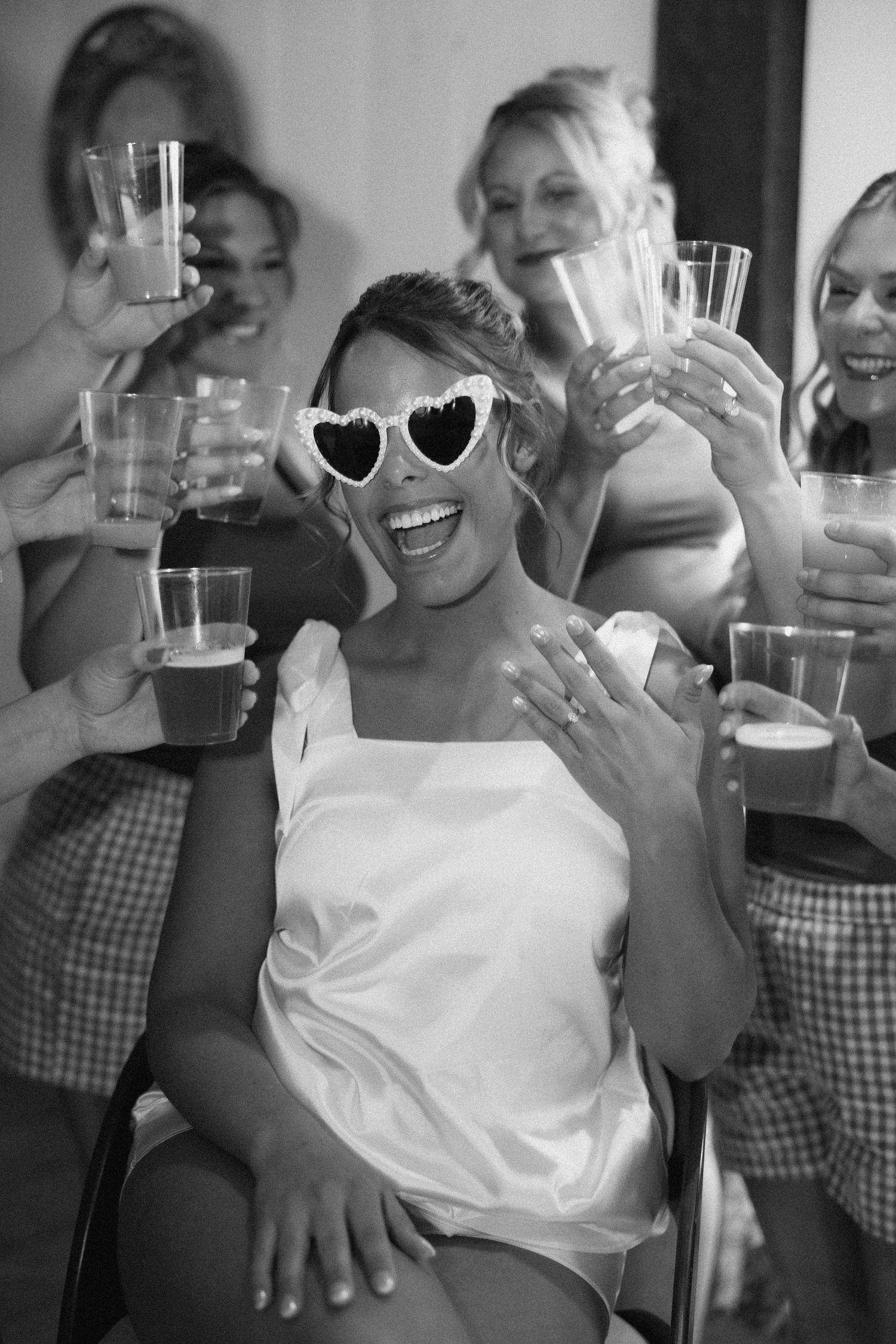 Laughing bride wearing heart-shaped sunglasses shows engagement ring at bachelorette party as friends raise glasses in a toast.