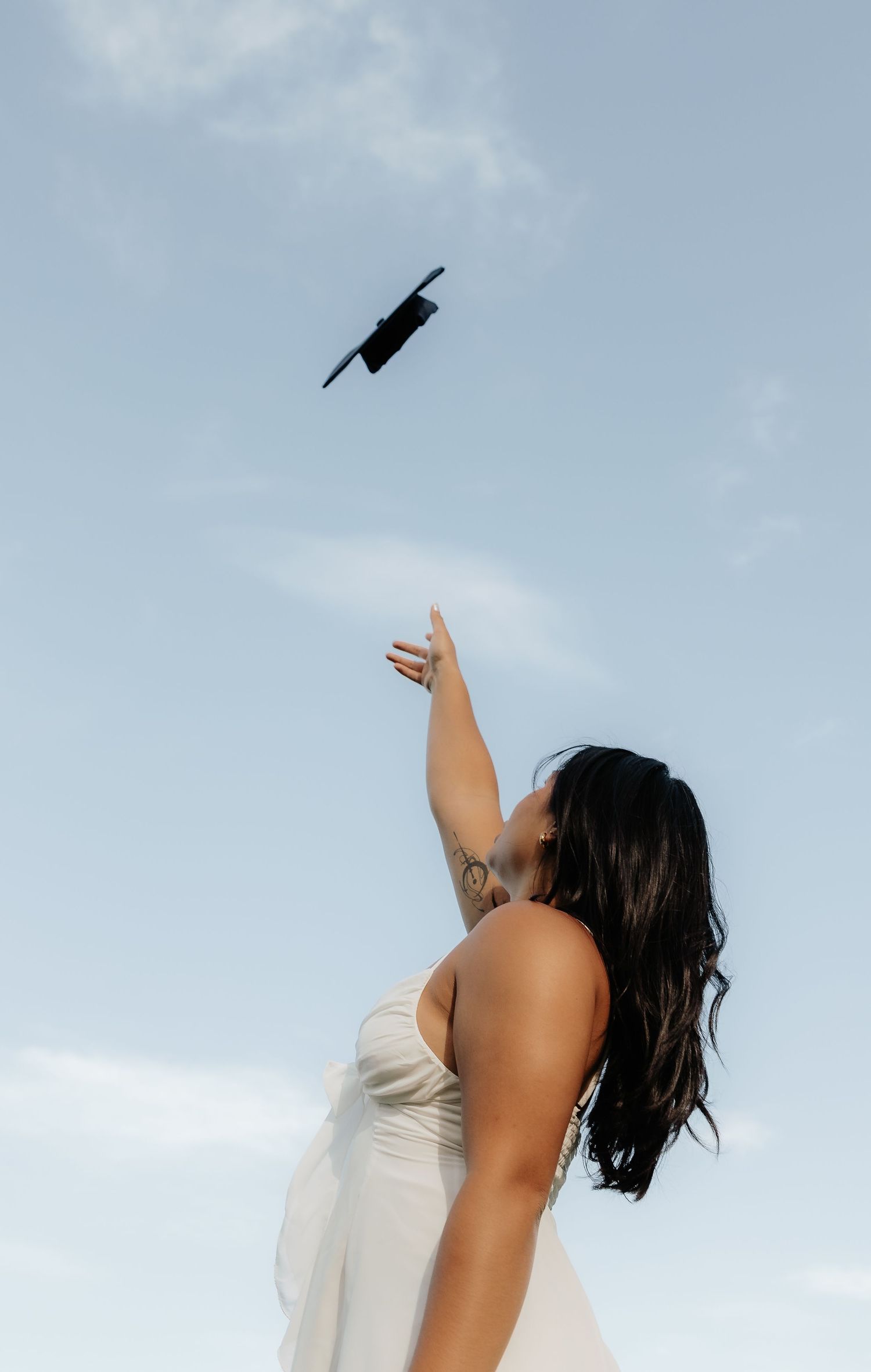 Woman in white dress tossing graduation cap into blue sky, celebrating academic achievement outdoors.