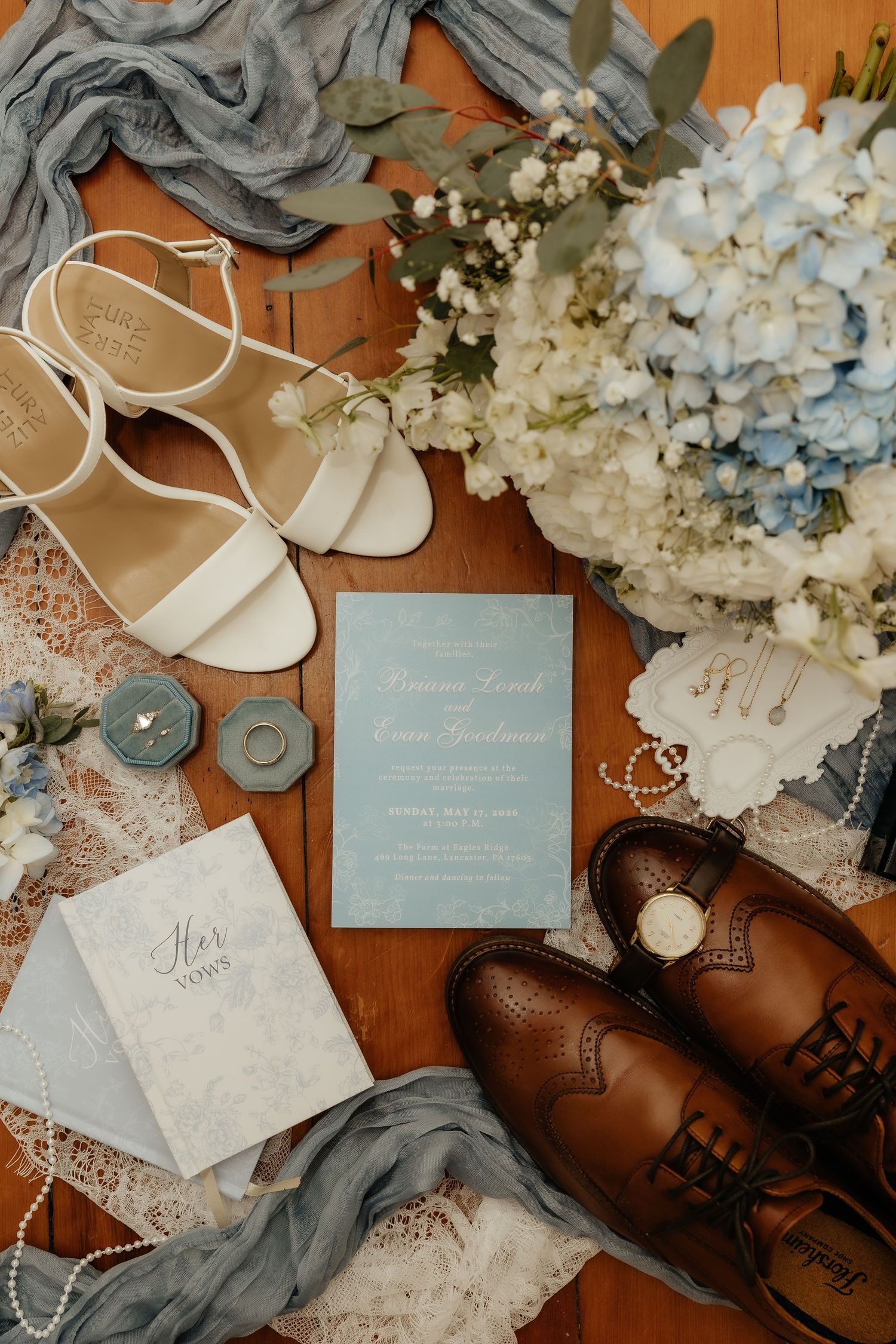 Elegant wedding flat lay with blue hydrangea bouquet, white heels, brown oxfords, ring boxes, pearl necklace, and blue invitation.