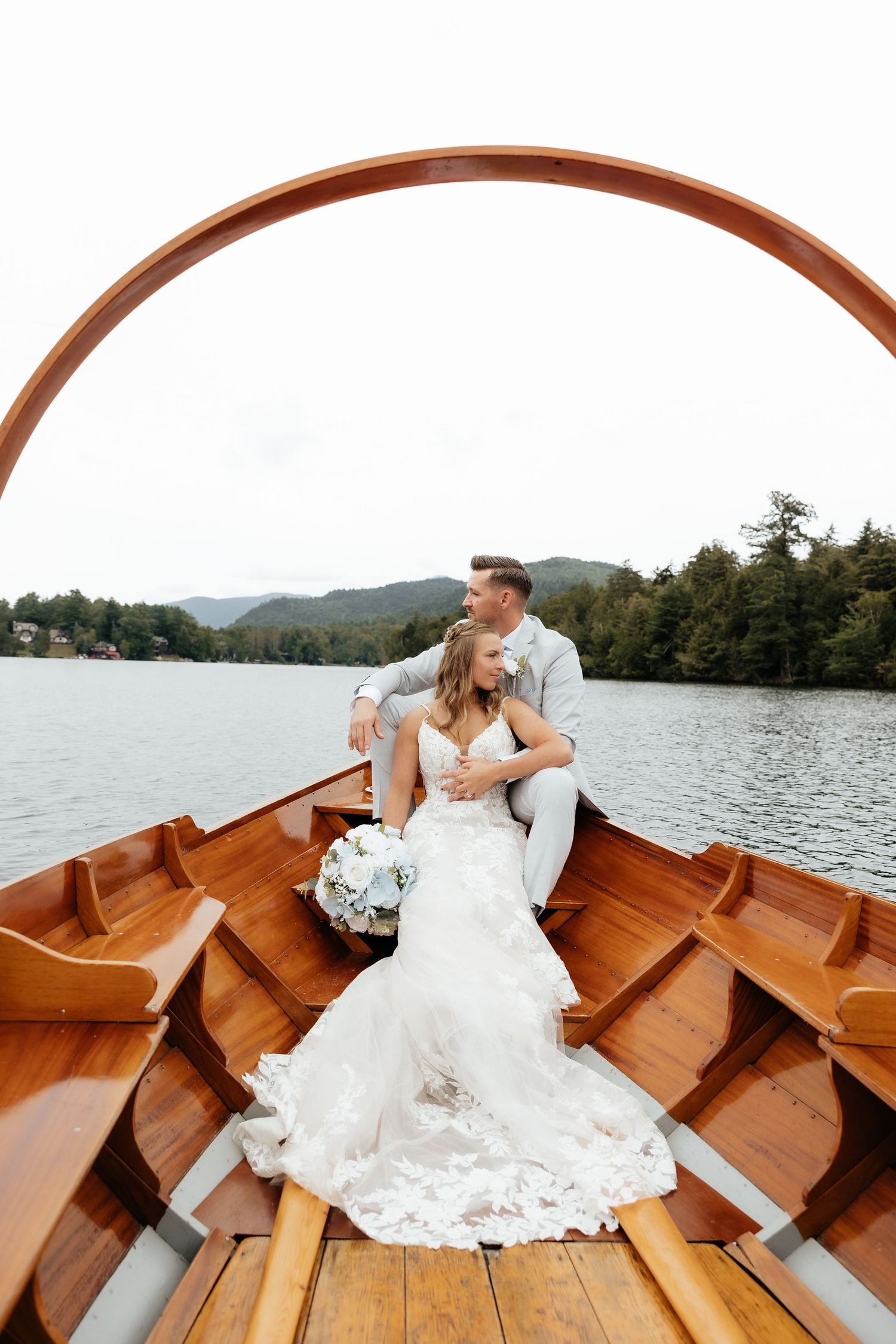 Bride and groom sitting together in a wooden rowboat on a scenic lake, bride holding white bouquet in wedding dress.