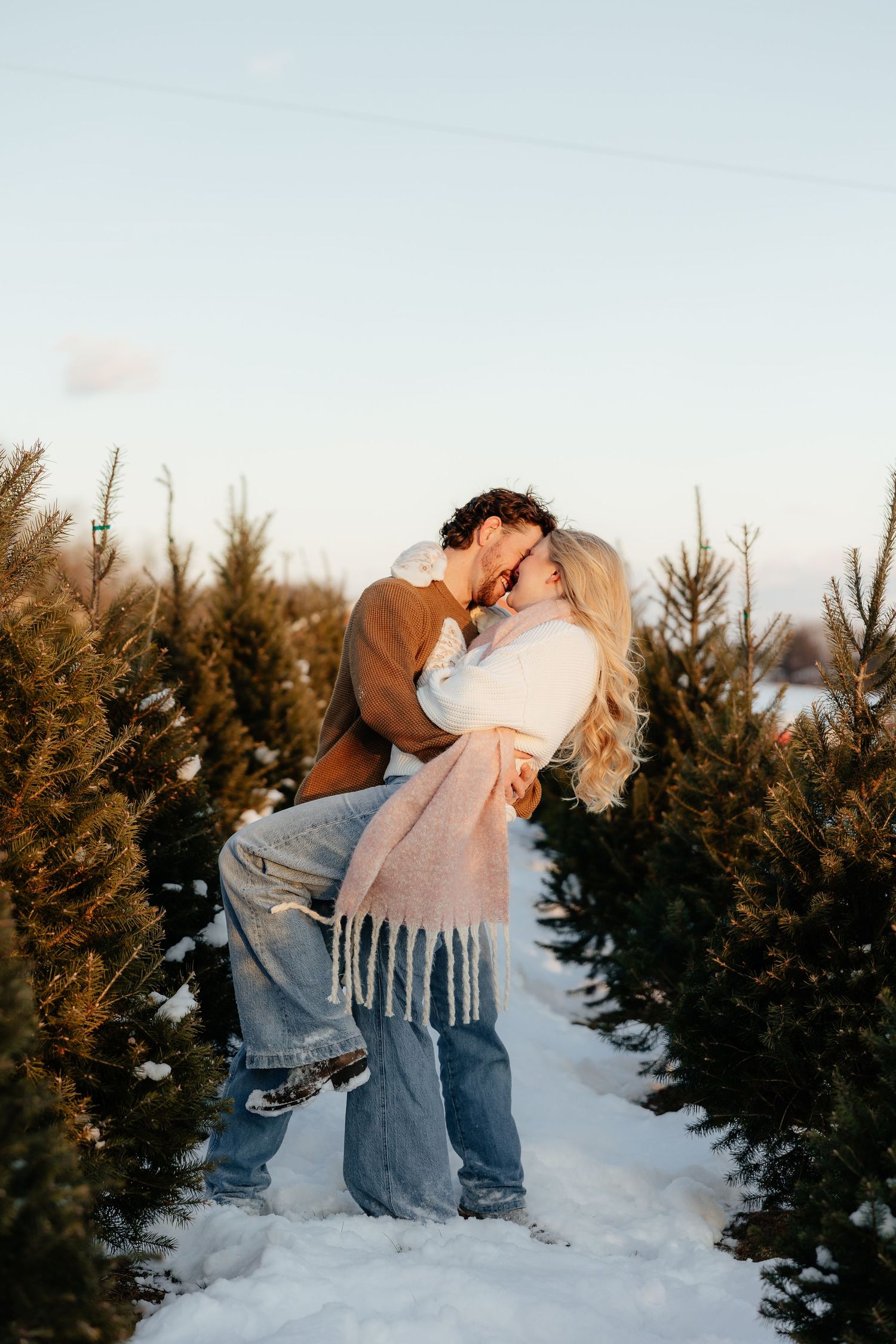A couple shares a romantic kiss in the snow at a Christmas tree farm during winter.