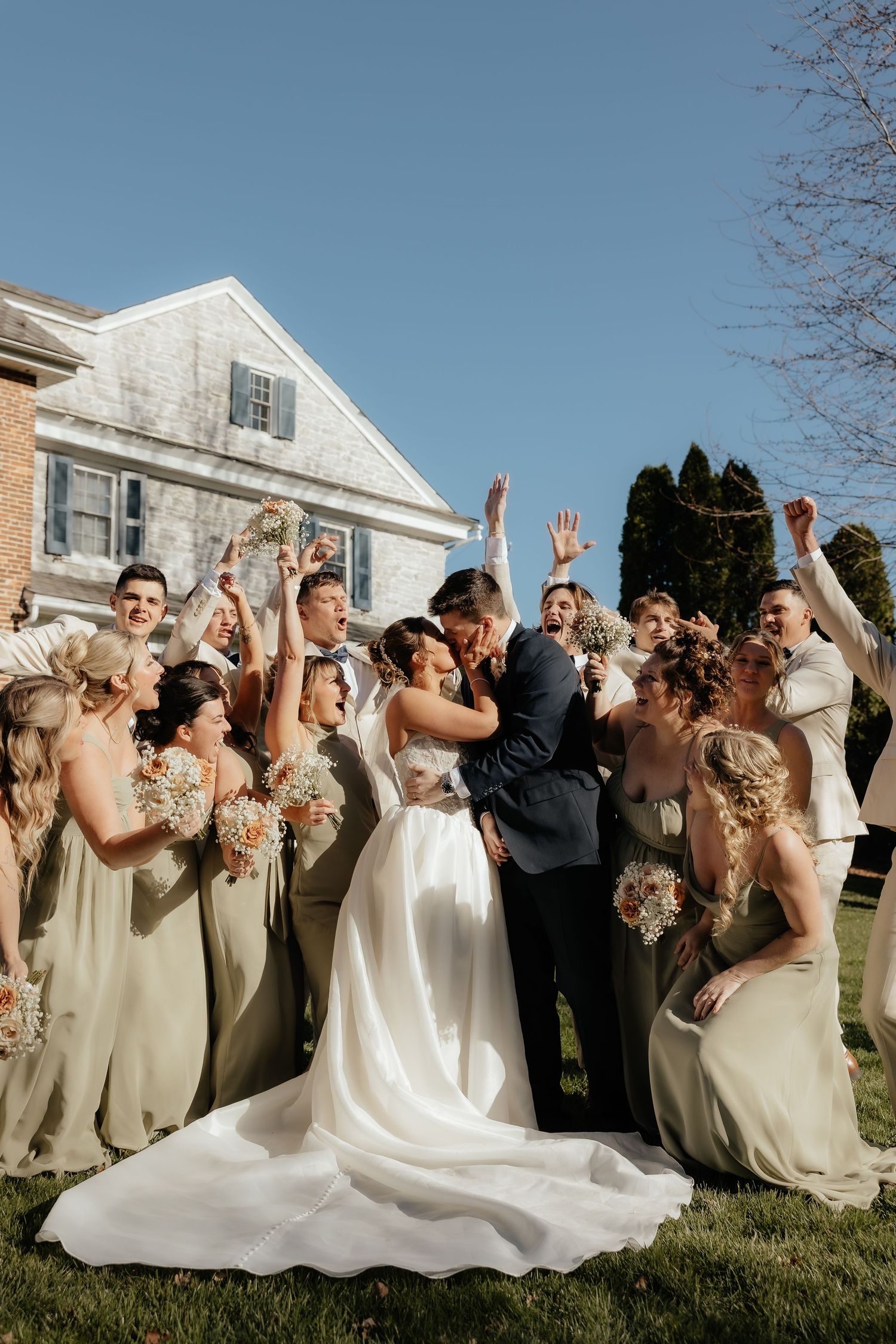 A bride and groom share a kiss surrounded by cheering bridesmaids in sage gowns and groomsmen outdoors on a sunny day.