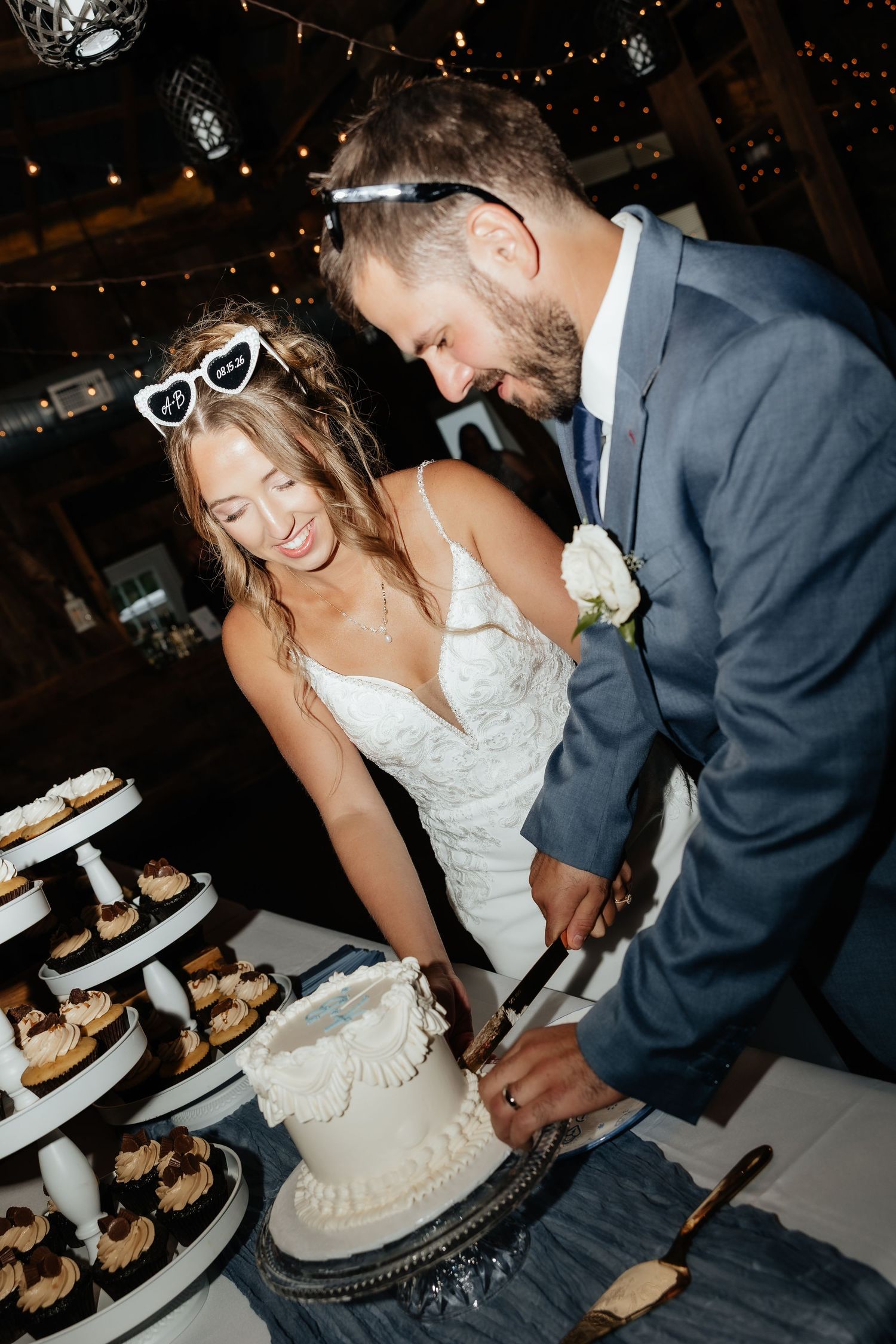 Bride and groom smiling while cutting their white wedding cake at a rustic barn reception.