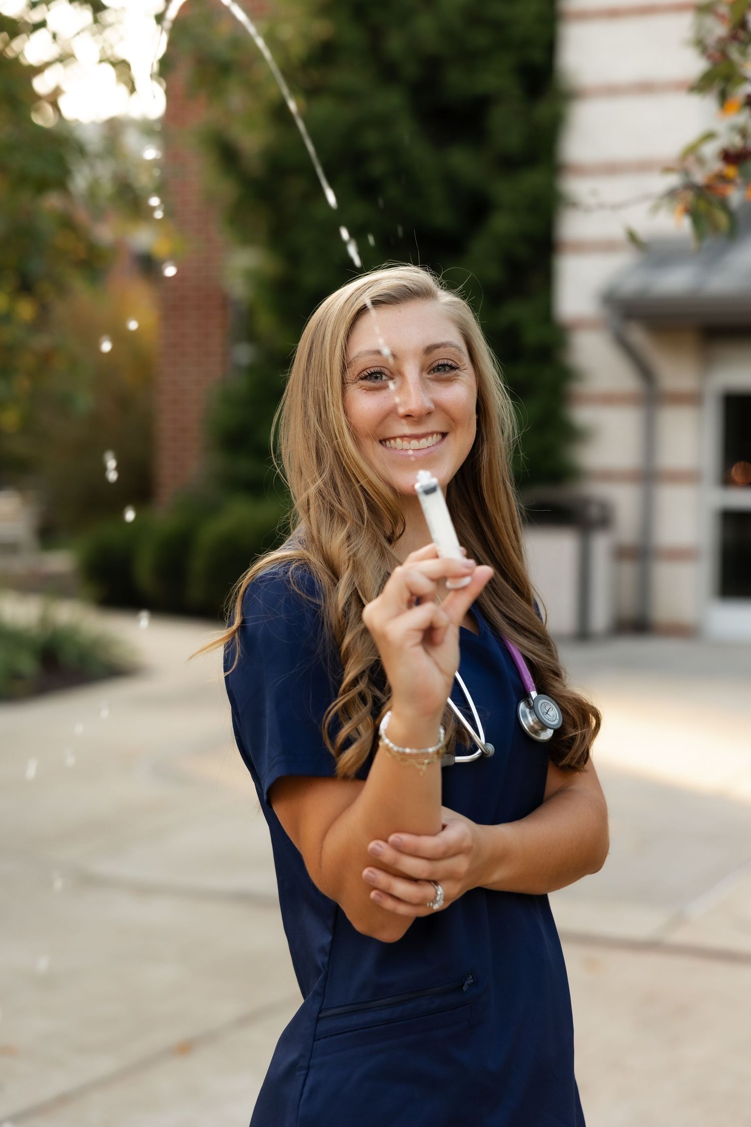 Healthcare professional in navy blue scrubs holds a stethoscope while standing outside a medical building.