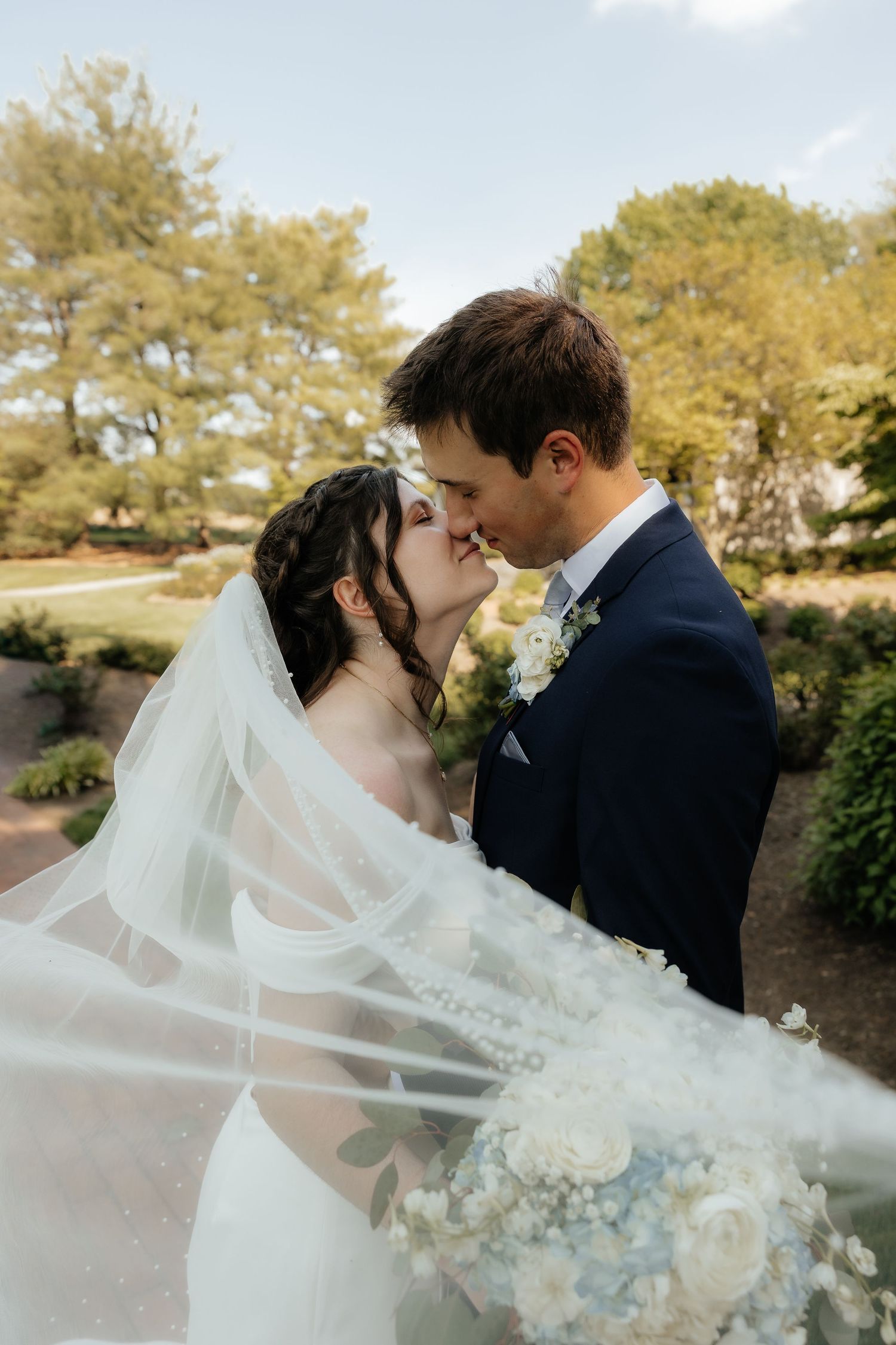 Bride and groom share a romantic kiss outdoors, with flowing veil and white floral bouquet, surrounded by lush greenery.