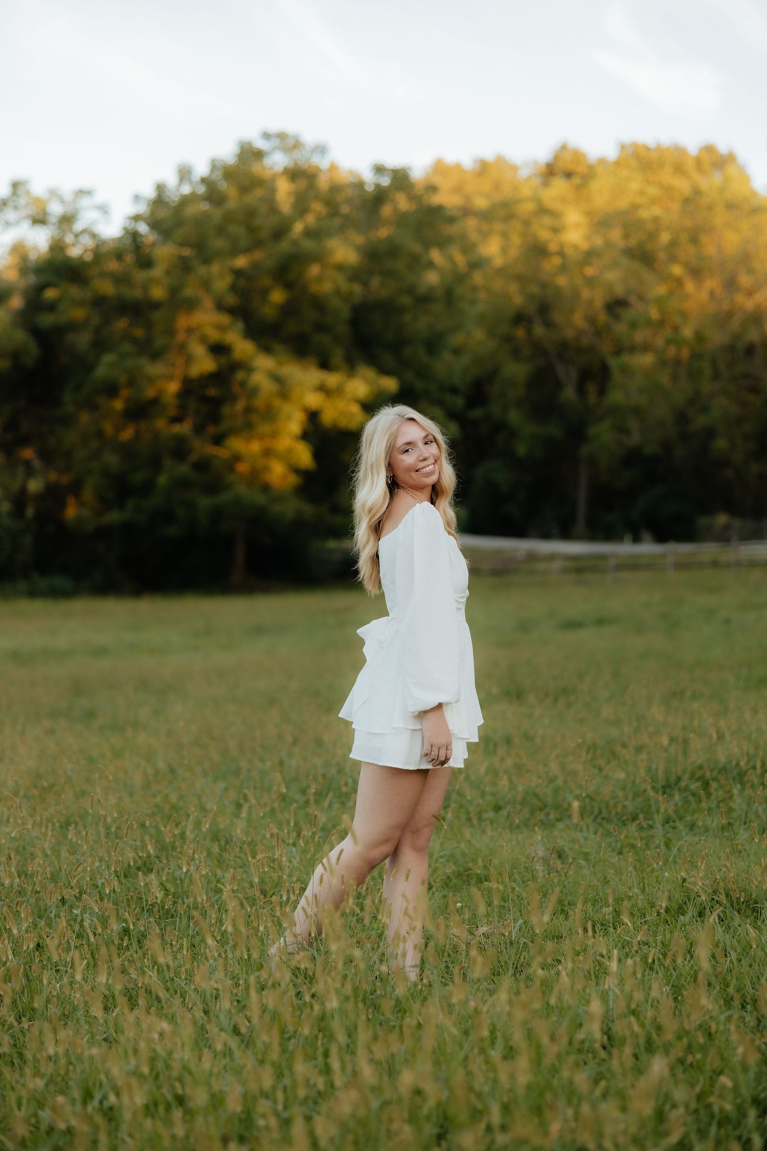 A person in a white dress twirls joyfully in a sunlit meadow with golden autumn trees in the background.