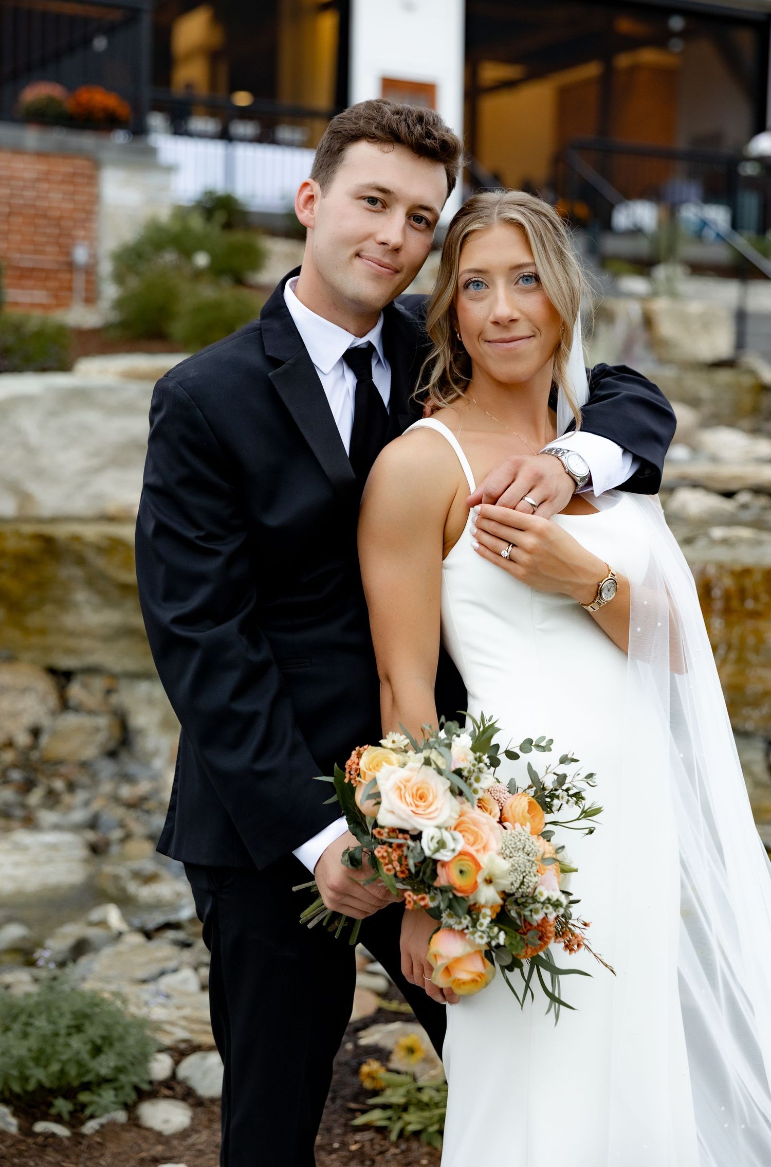 A romantic wedding couple embraces outdoors, with the bride holding a peach and white floral bouquet.