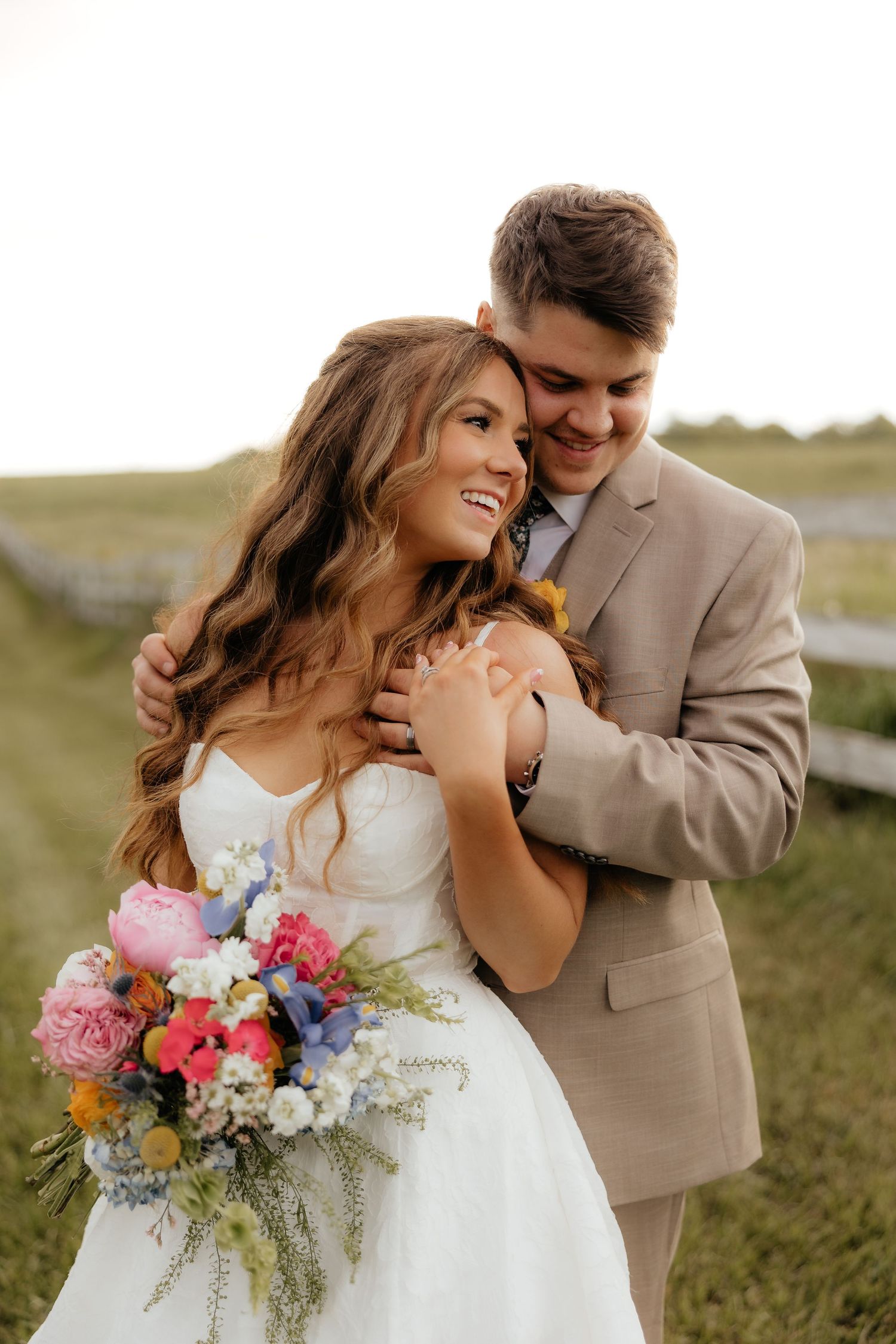 Bride and groom embrace outdoors, she holds colorful bouquet in white dress, he wears tan suit, smiling joyfully.