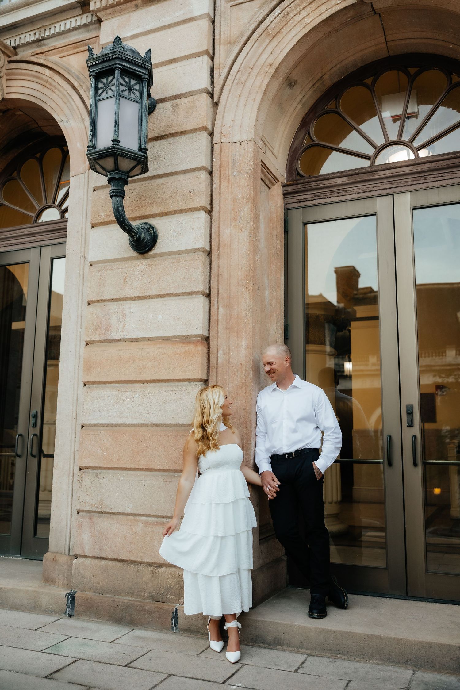Couple in wedding attire holding hands outside elegant stone building with ornate lantern and arched doors.