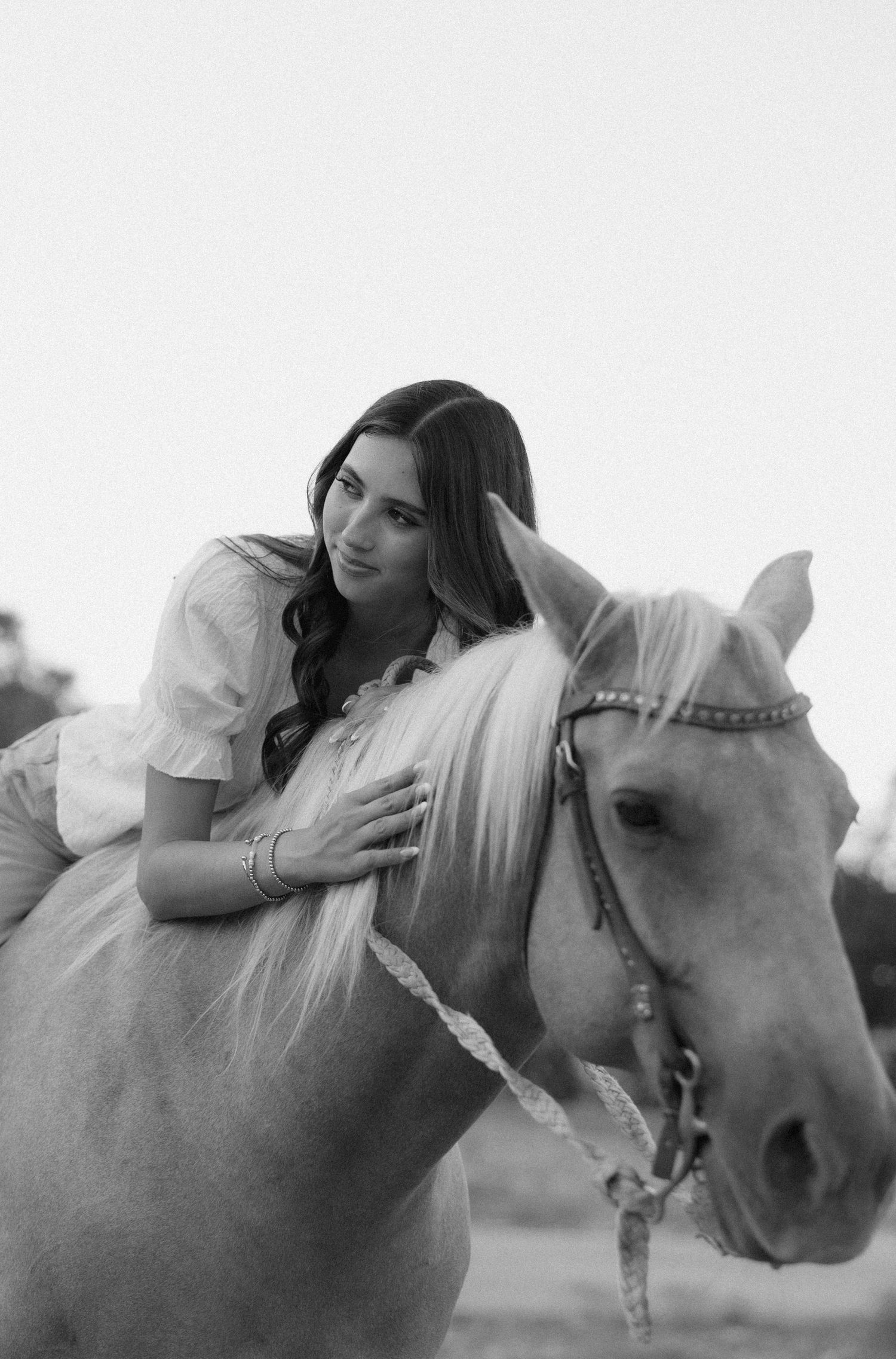 Black and white photo of a young woman in a white dress leaning affectionately on a light-colored horse with a decorative bridle.