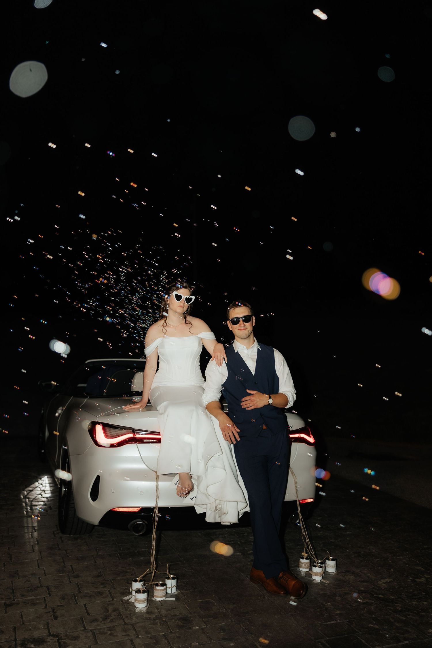 Bride and groom in sunglasses pose against white convertible at night with confetti falling around them.
