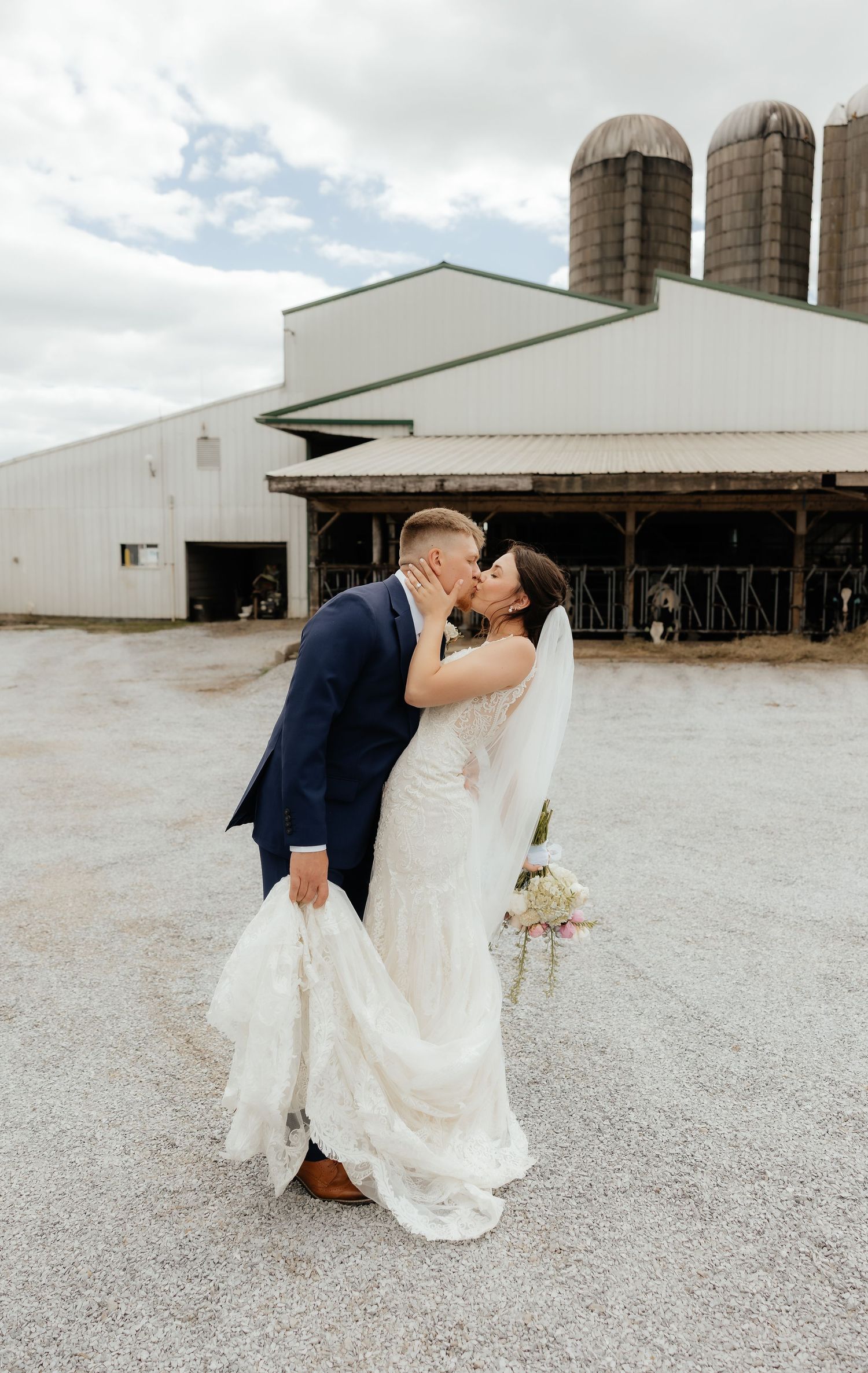 Bride and groom share a romantic kiss at a rustic farm wedding venue with silos and barn in the background.