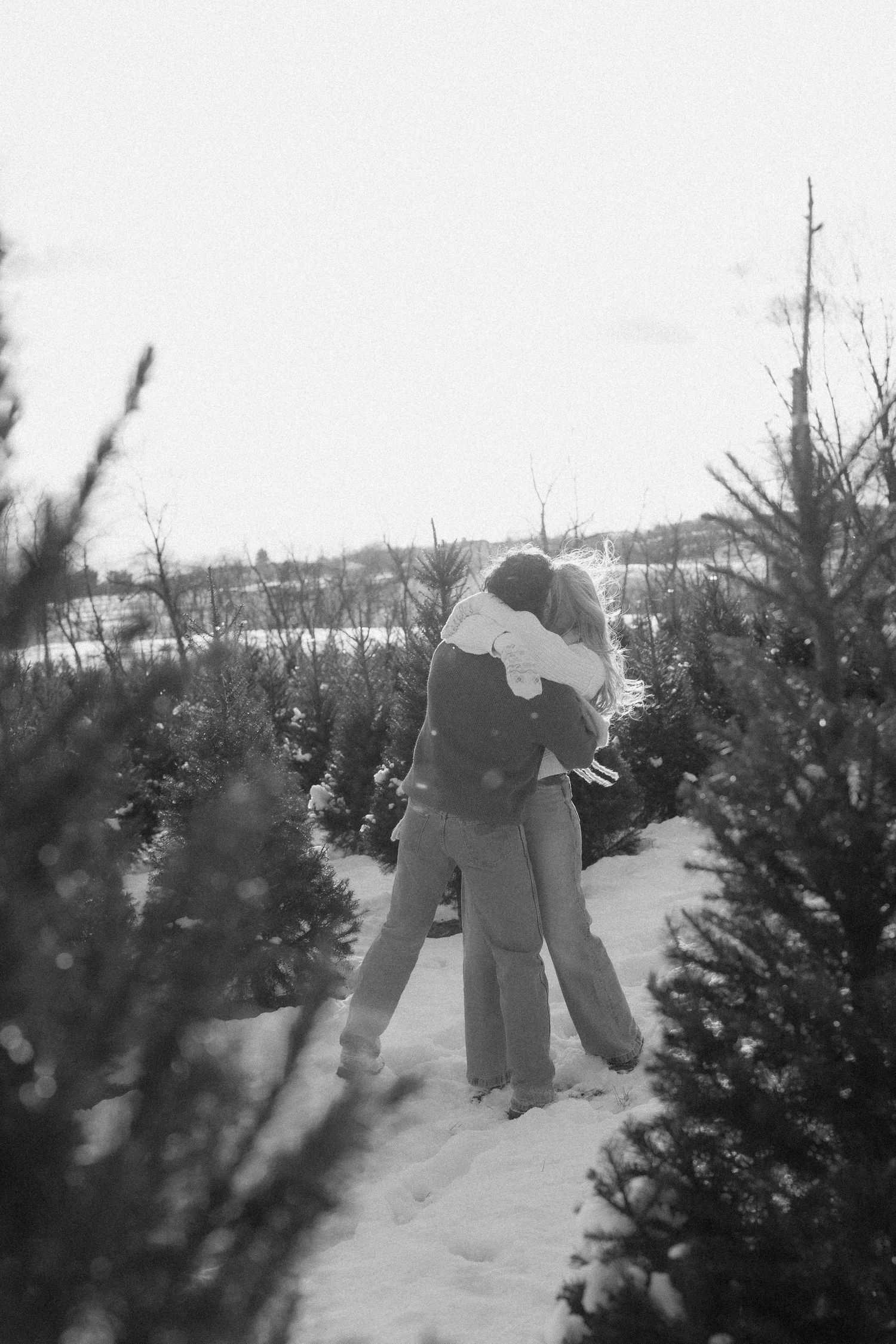 People browse rows of evergreen Christmas trees at a snowy outdoor tree lot during winter.