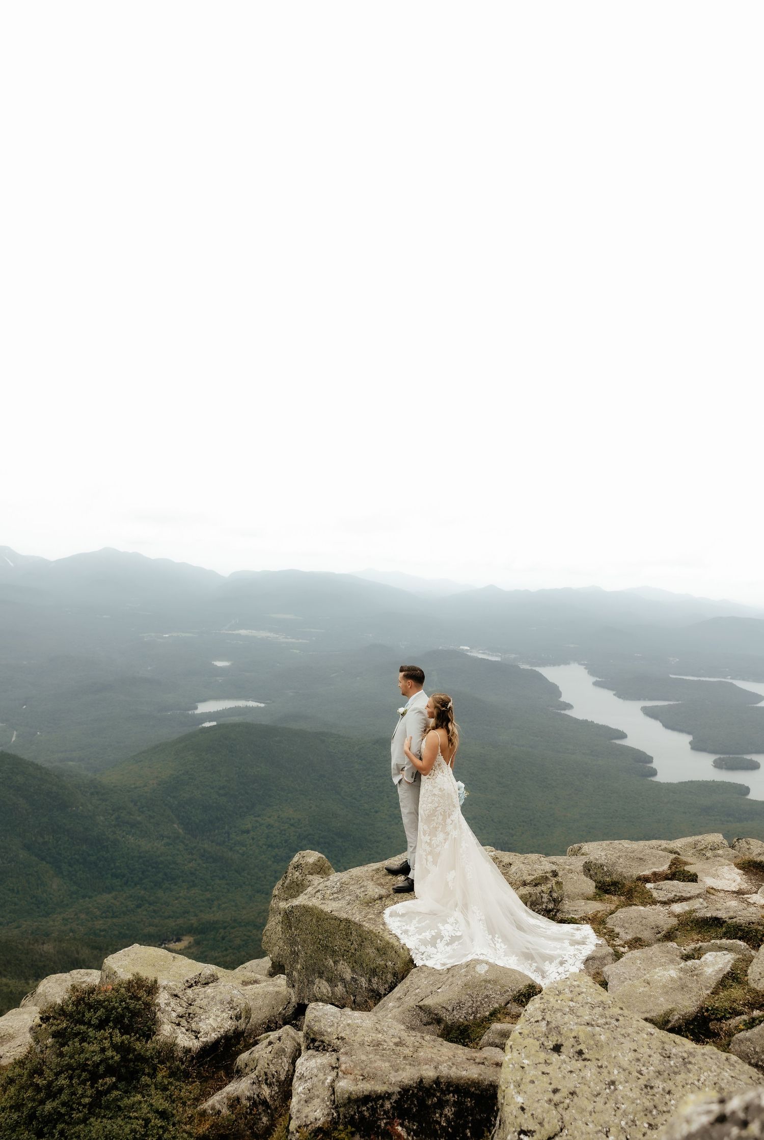 Bride and groom stand on rocky mountain summit in wedding attire, overlooking lush green valleys and winding river below.
