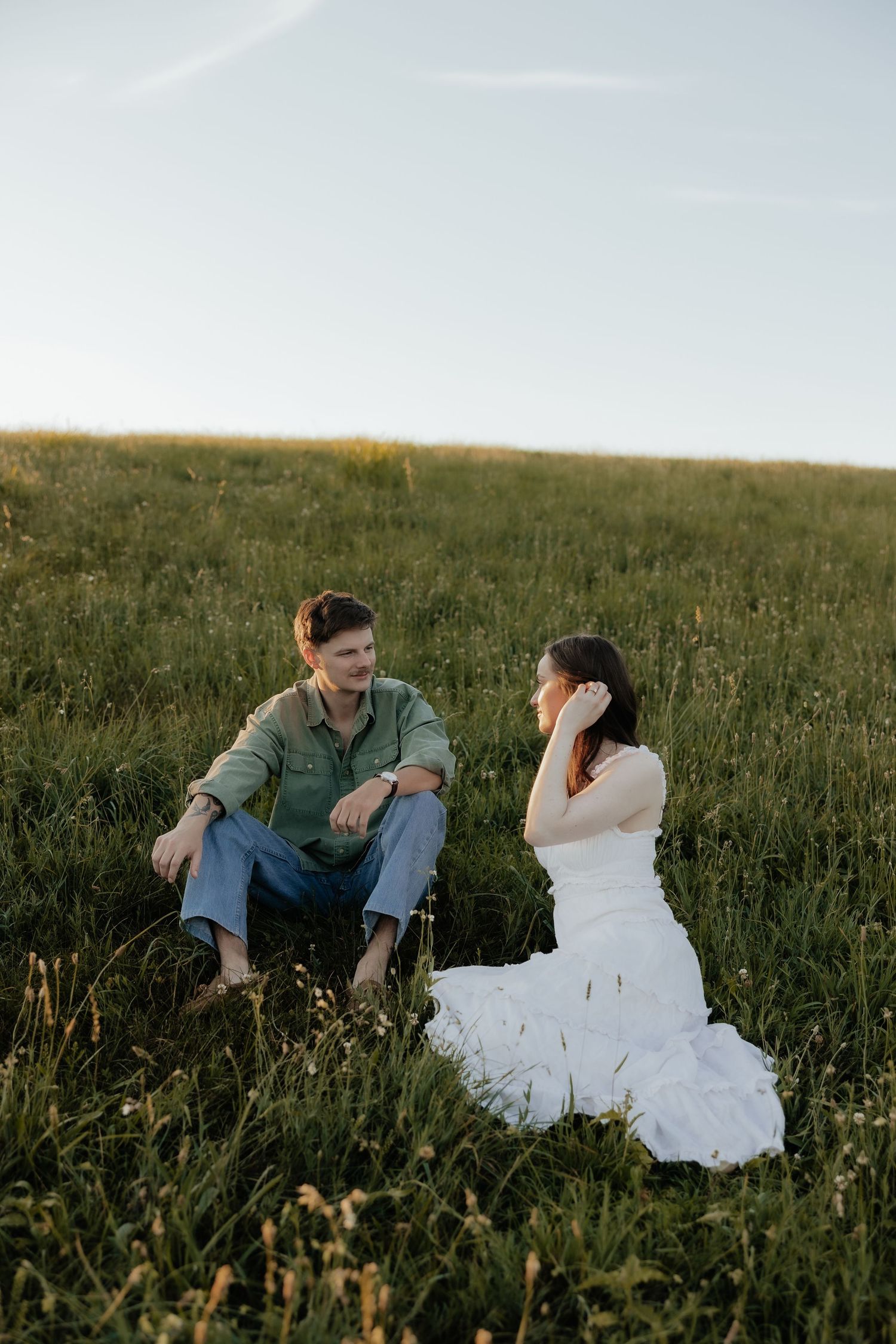 A bride in a white gown and groom in denim sit together on a lush green meadow during golden hour.
