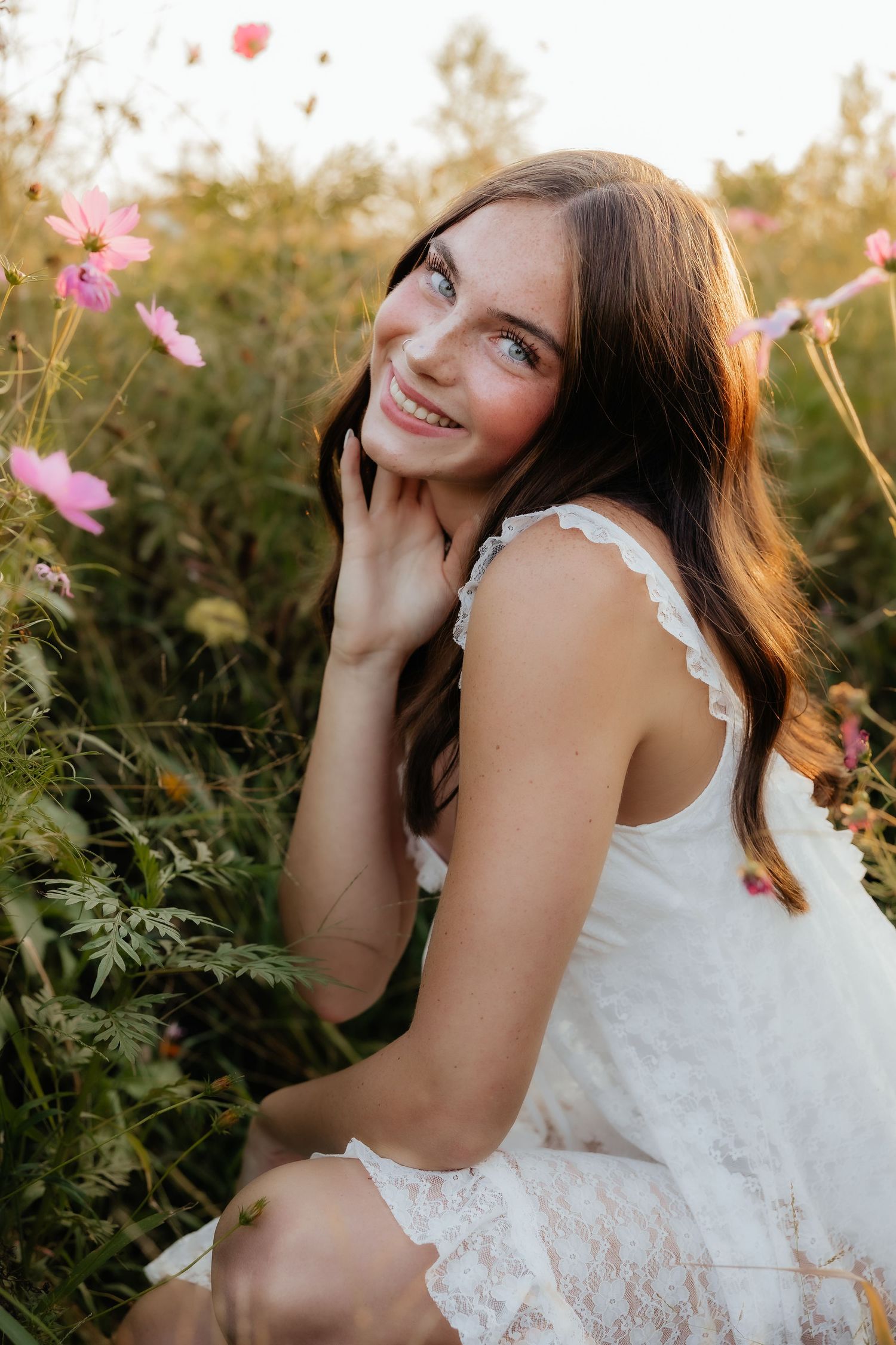 Young woman in white lace dress smiling in a field of pink wildflowers during golden hour.