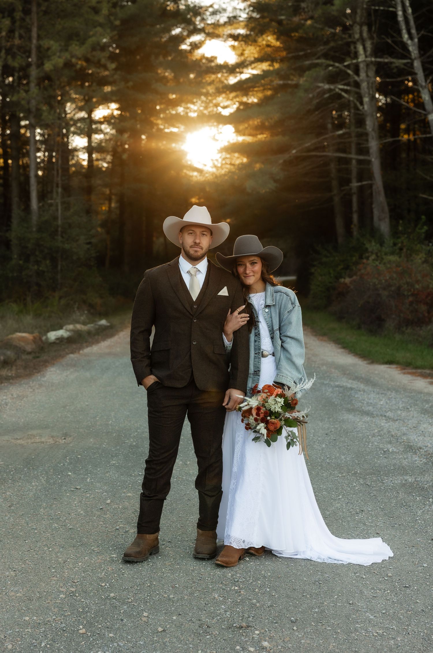 A couple in western attire poses on a dirt road at sunset with pine trees in the background.