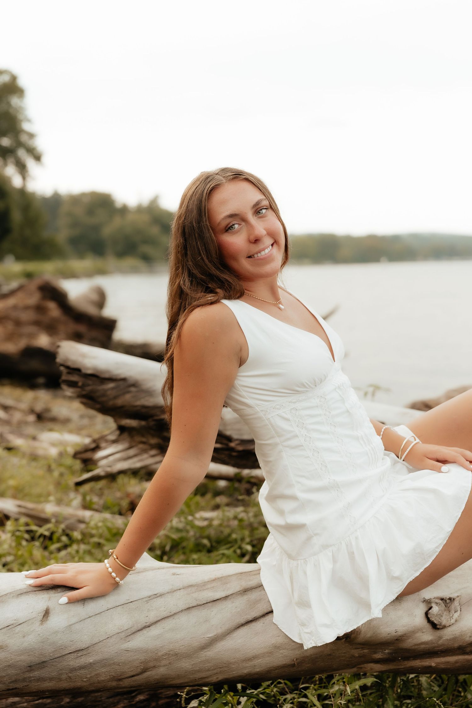 Young woman in white dress sitting on driftwood logs by a lake, smiling outdoors in a natural setting.
